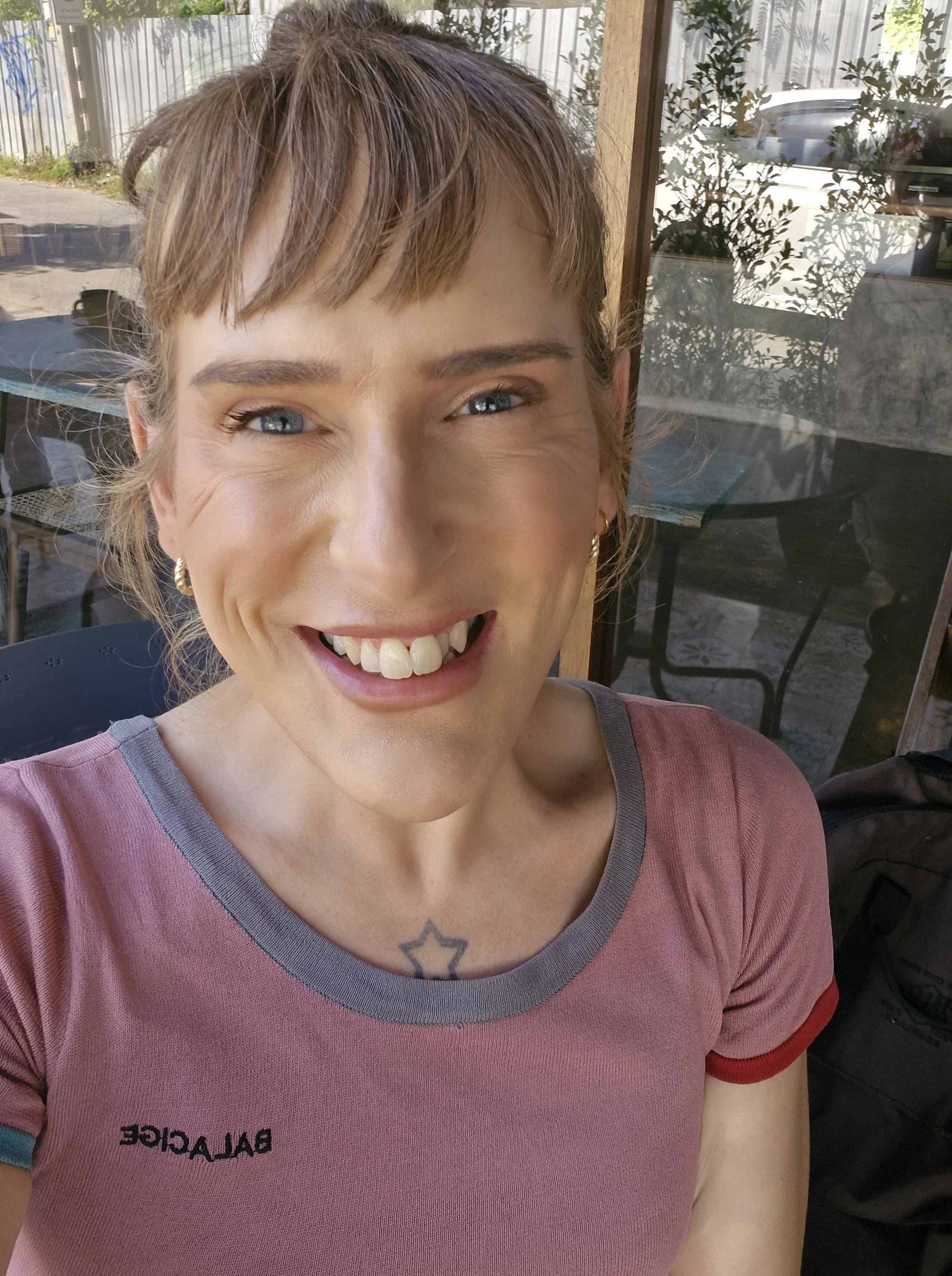 A woman with short, light brown hair and blue eyes smiling broadly indoors, wearing a pink shirt with gray collar and a small black star tattoo on her chest, with a reflection of trees and outdoor scenery in the window behind her.