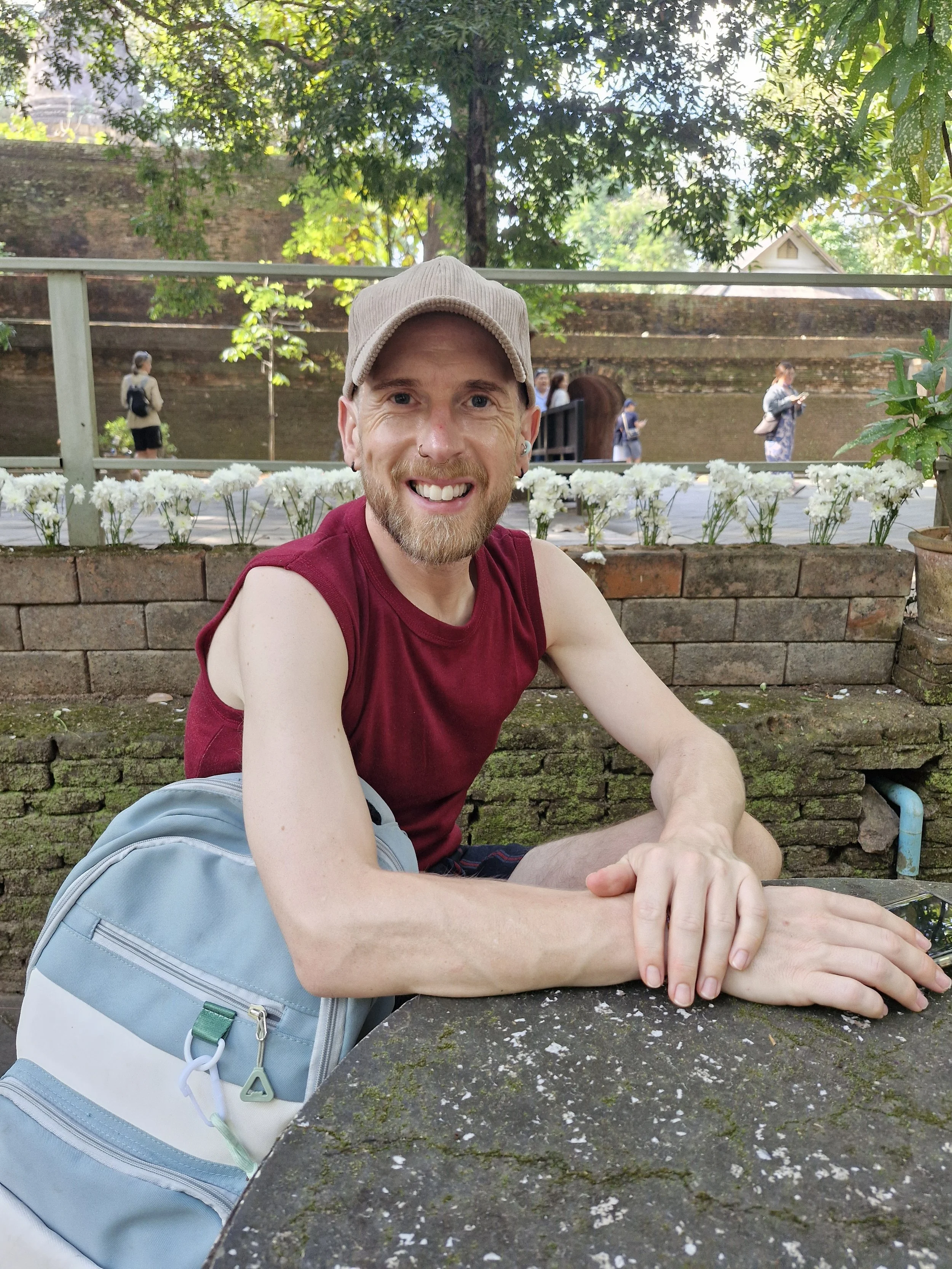 A smiling person , wearing a beige cap and a sleeveless maroon shirt, sitting outdoors. Behind him, there is a brick wall and a display of white flowers, with several people walking along a path in a park or garden setting under green trees.