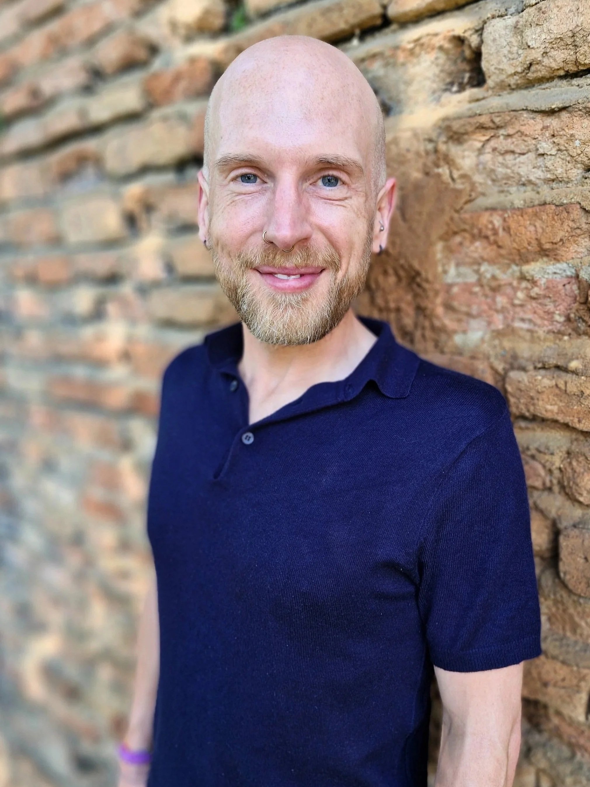 A smiling non-binary person with a shaved head, blue eyes, and a beard, wearing a black polo shirt, standing against a brick wall.