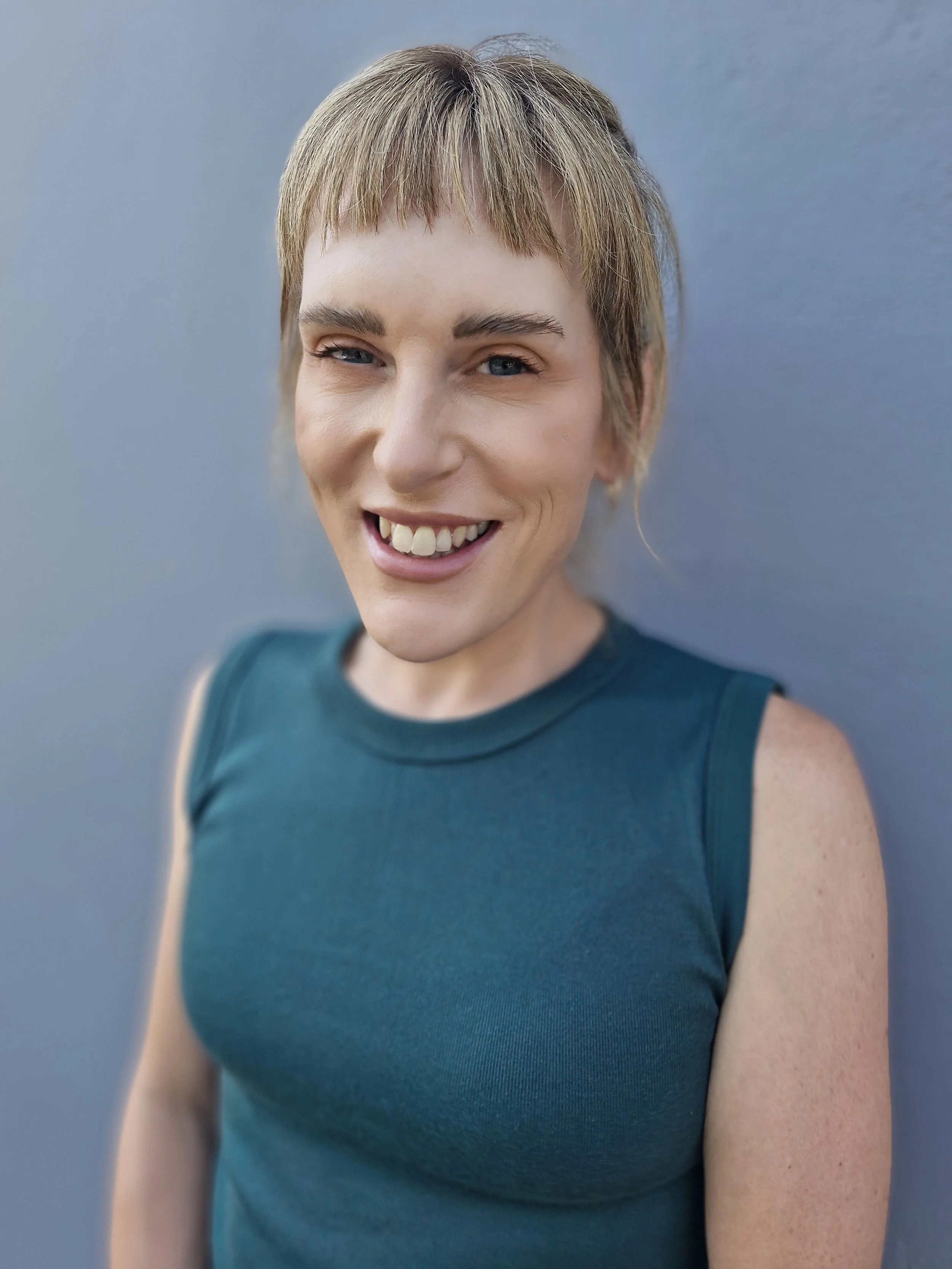 Close-up of a smiling woman with short blonde hair, wearing a sleeveless teal top, standing against a plain blue-gray background.
