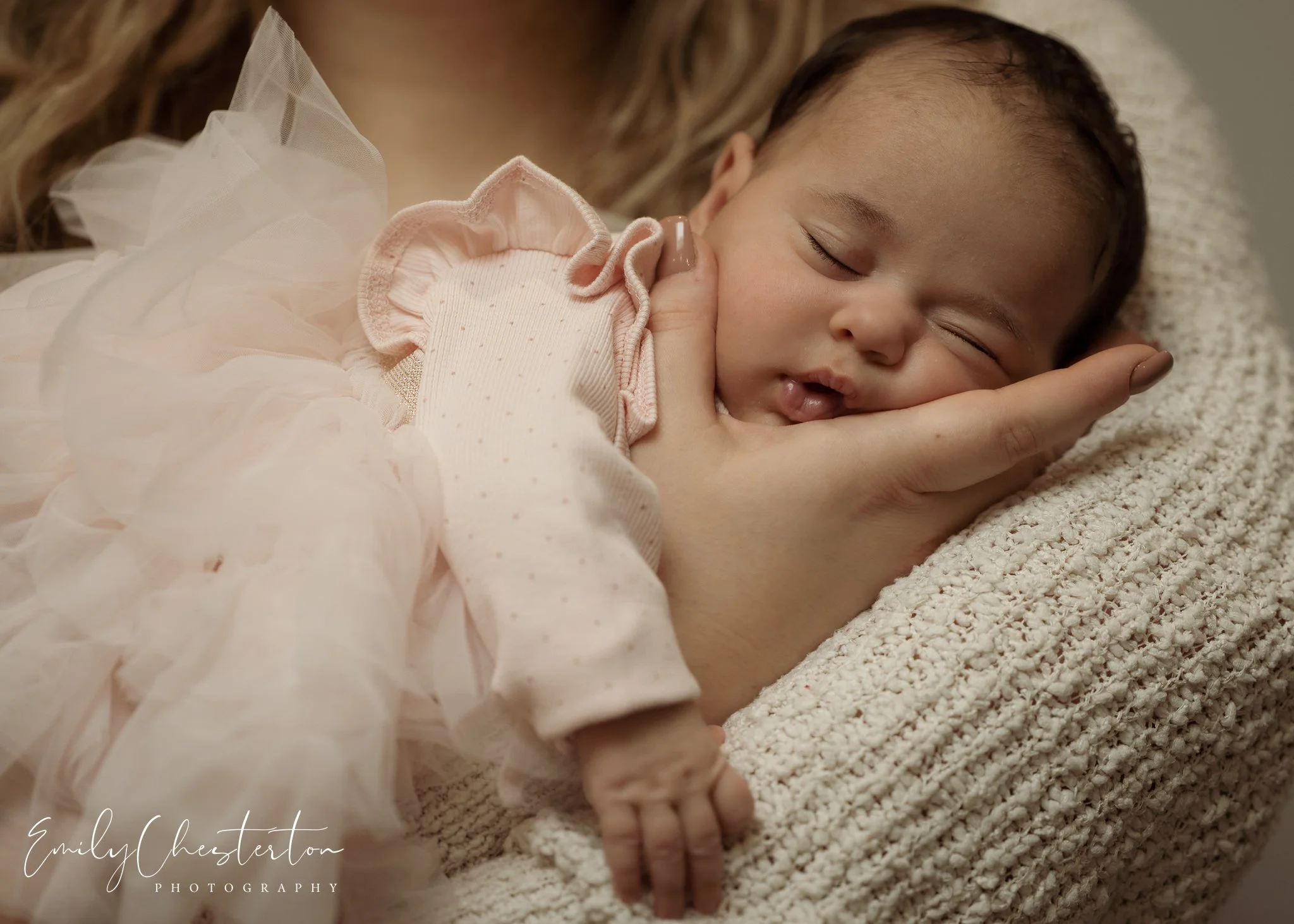 A sleeping infant being gently held by an adult's hand, resting on a fluffy blanket, dressed in a light pink dress with ruffled sleeves.