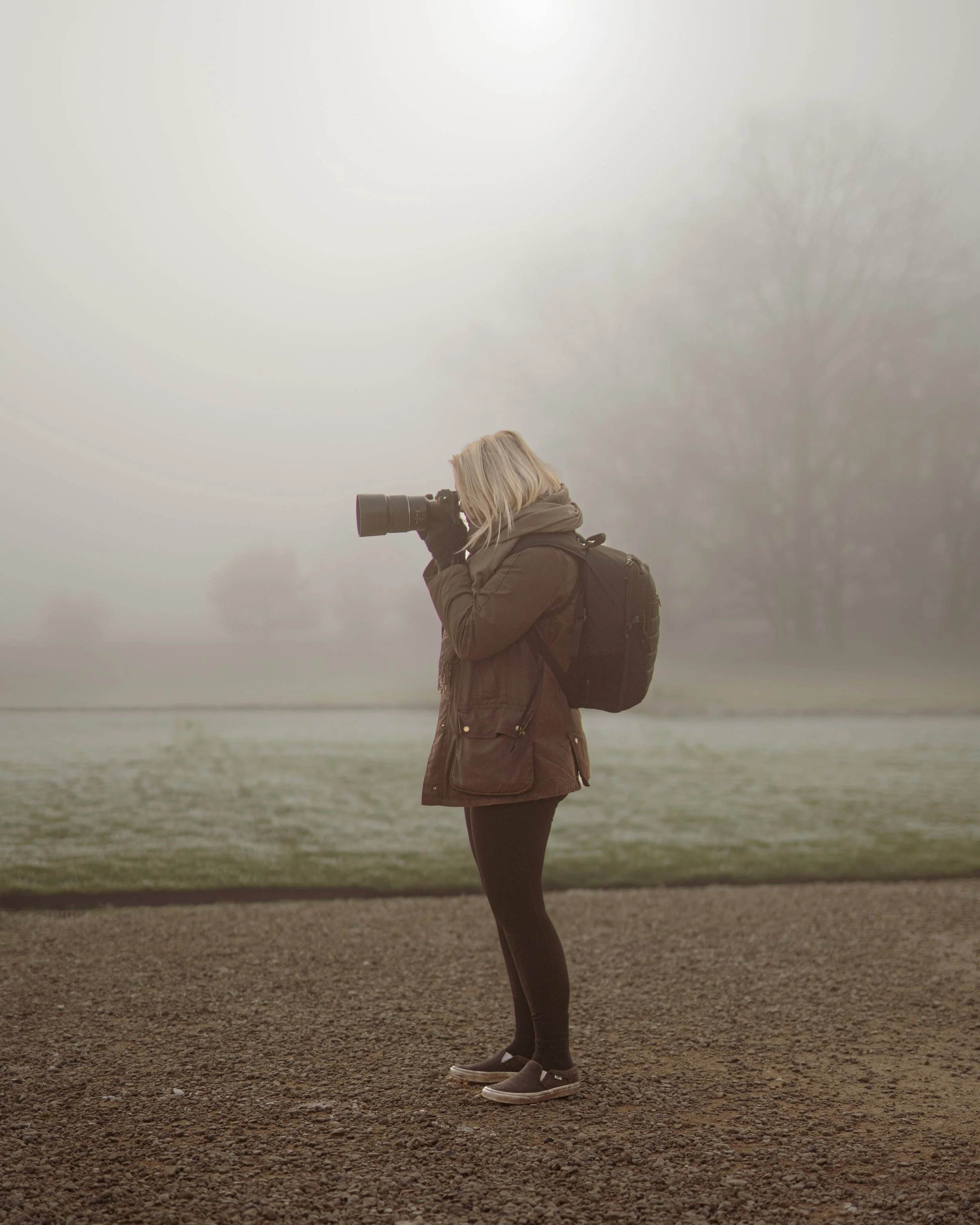 A woman standing outdoors on a foggy day, taking a photograph with a camera. She is wearing a brown coat, black pants, ankle boots, and has a black backpack.