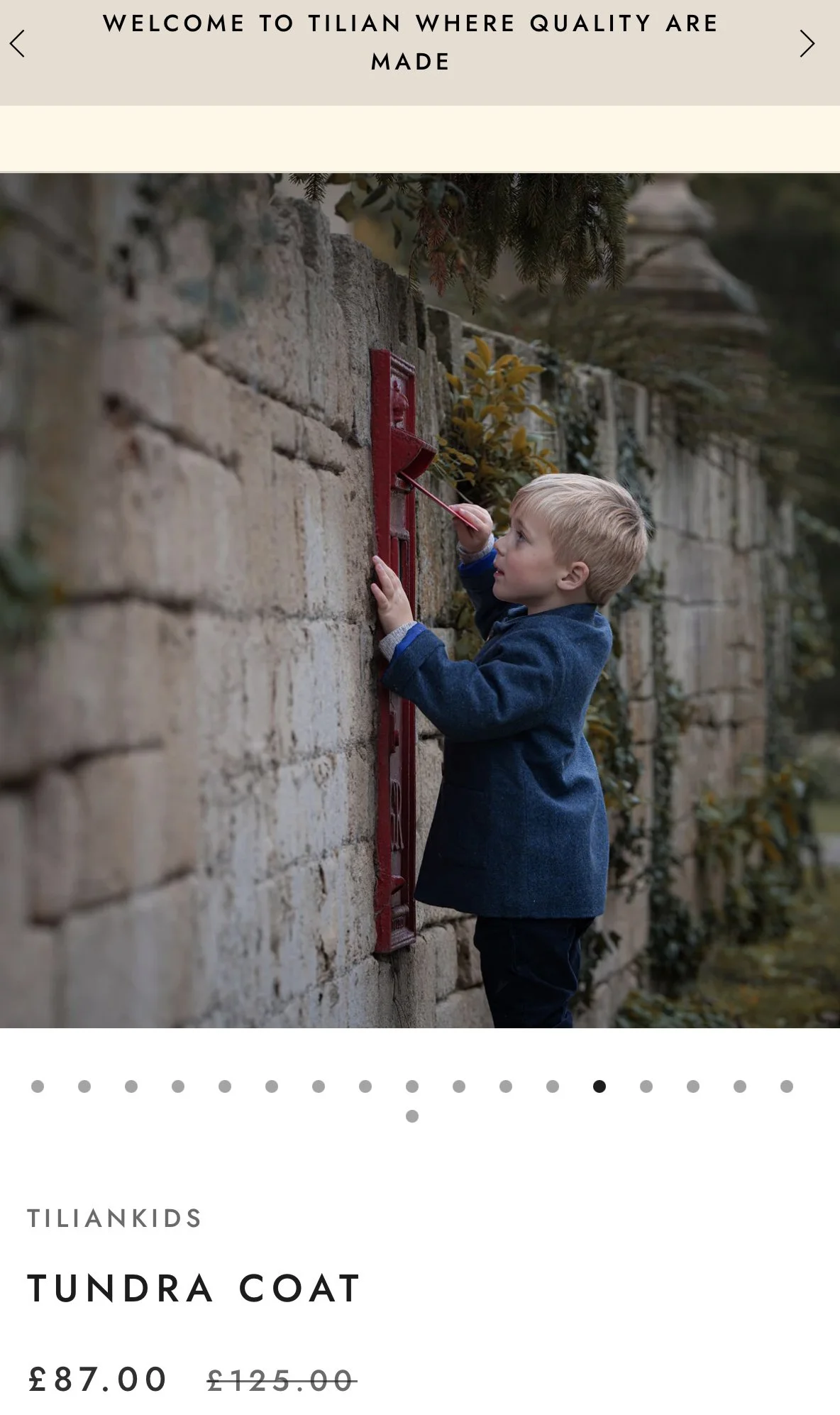 A young boy in a blue coat is painting a red letterbox on a stone wall. There are plants and trees in the background.