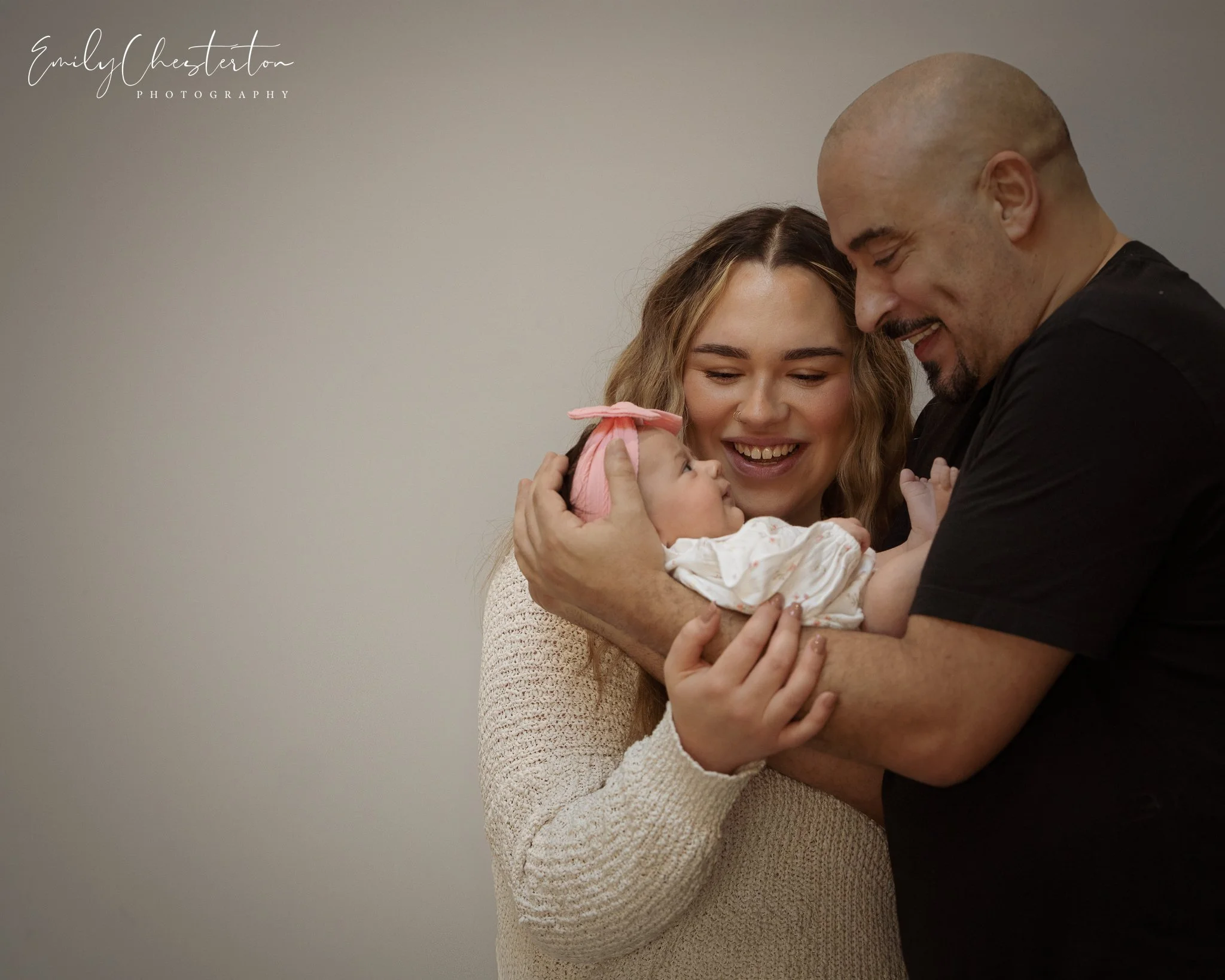 A woman and man holding a newborn baby girl, all smiling and looking at each other, against a plain beige background.