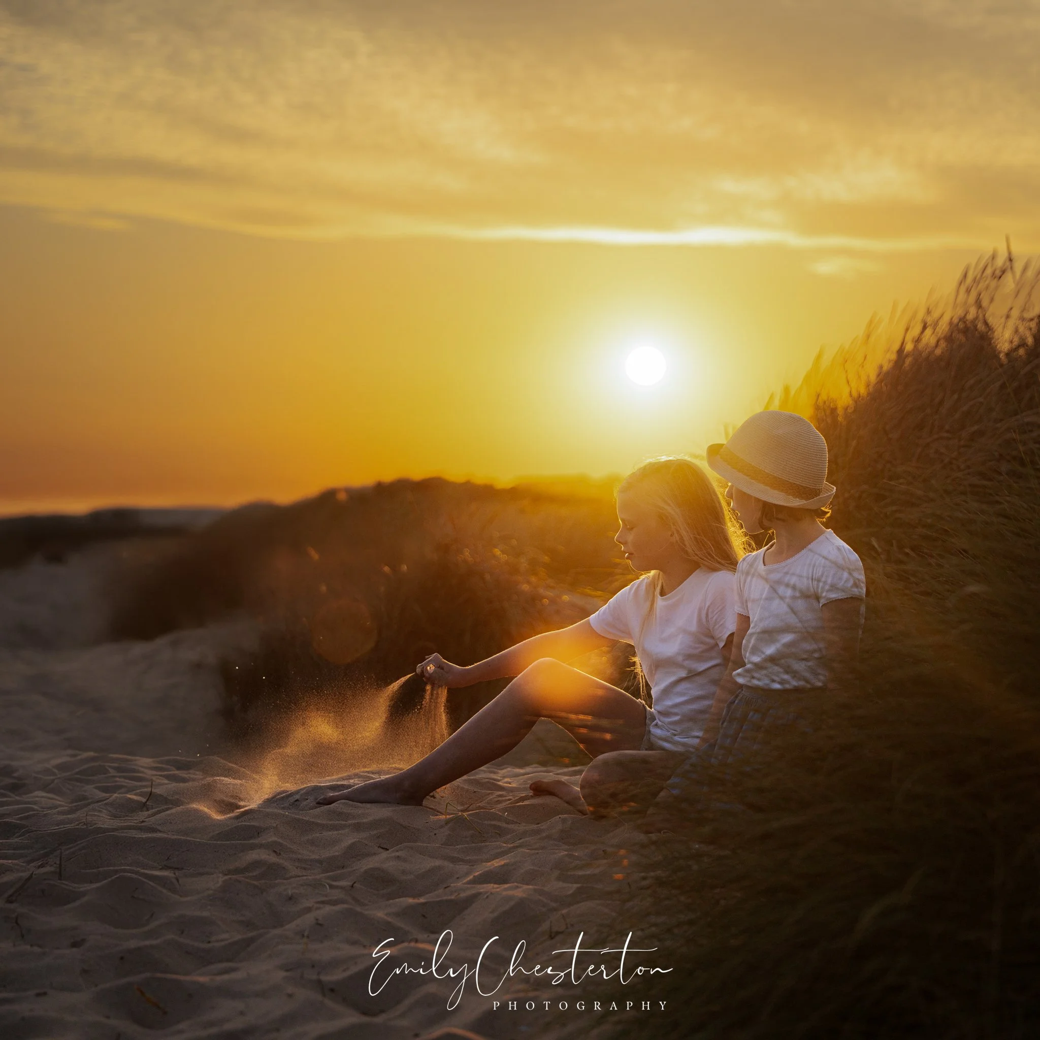 Two young girls sitting on a sandy beach at sunset, with one girl reaching down to touch the sand and the other girl wearing a sun hat, surrounded by beach grass and with the sun in the sky.