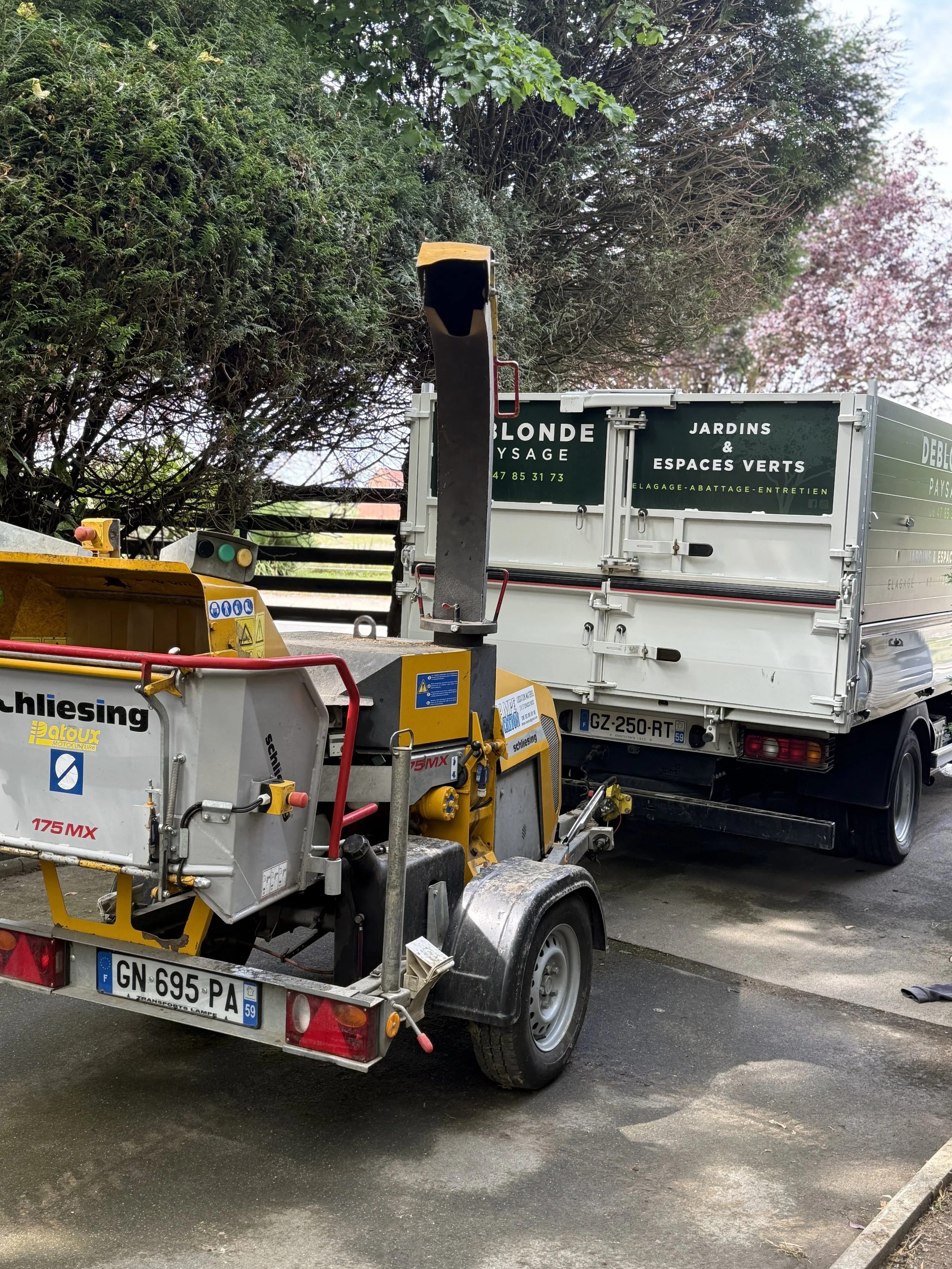 Un véhicule de travaux avec une machine en remorque, stationné devant un camion avec une inscription sur la porte arrière indiquant des services de jardinage et d'entretien paysager, dans un environnement extérieur avec des arbres en arrière-plan.