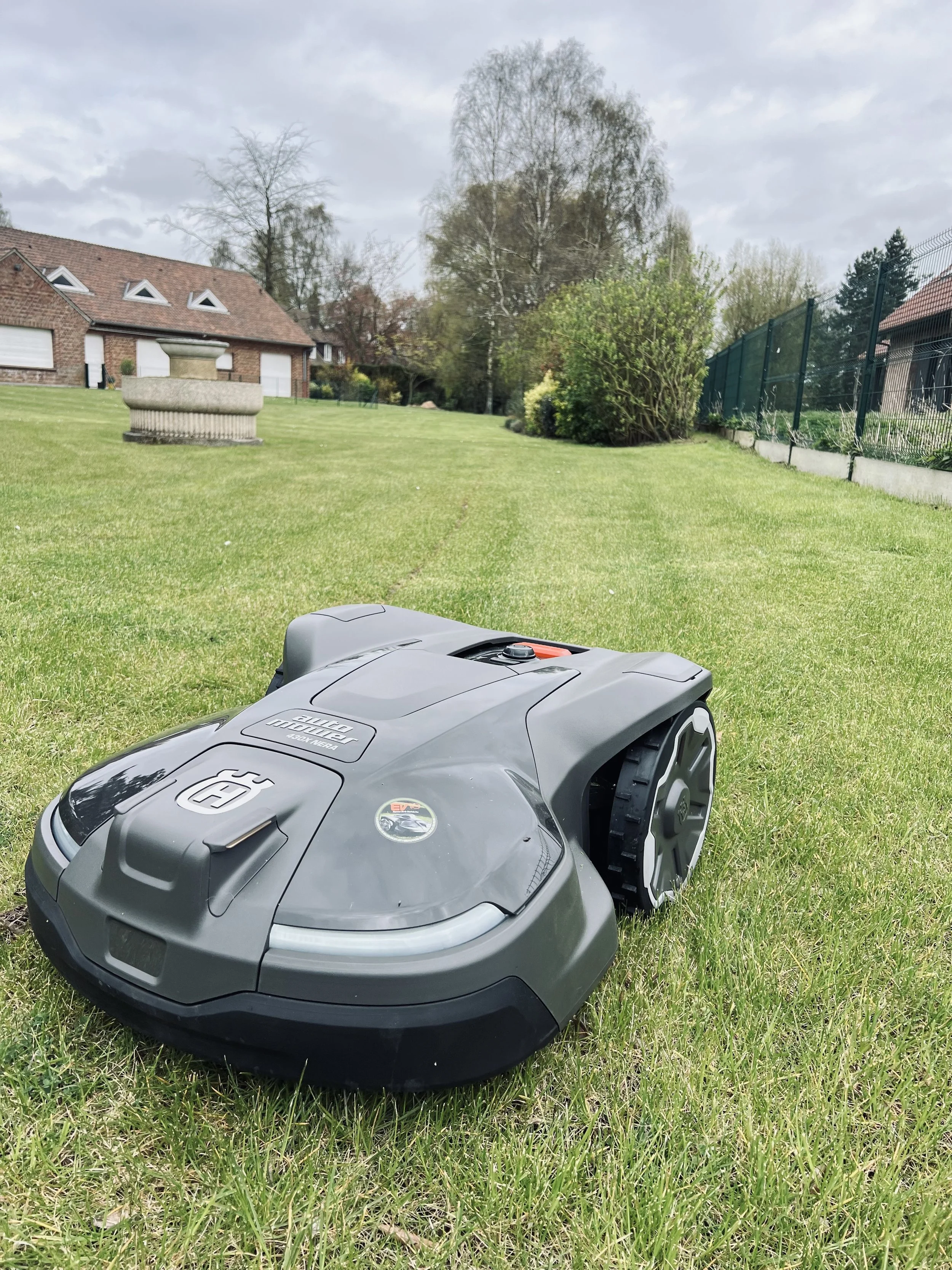 Un robot tondeuse sur une pelouse, avec des maisons et des arbres en arrière-plan sous un ciel nuageux.