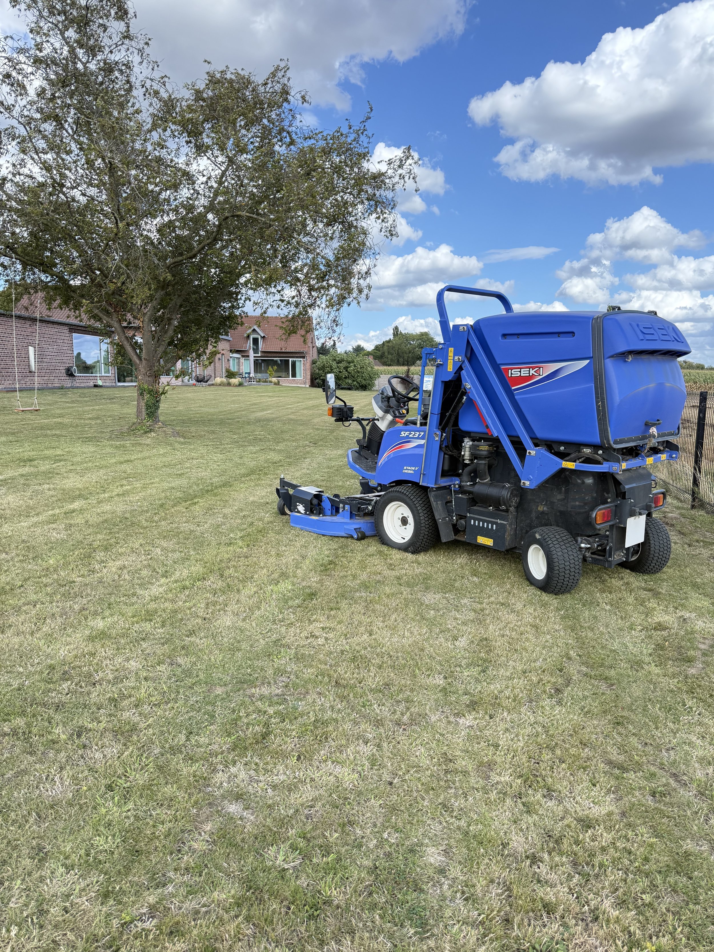 Une pelleteuse bleue stationnée sur une pelouse devant une maison avec un arbre en face et un ciel partiellement nuageux.
