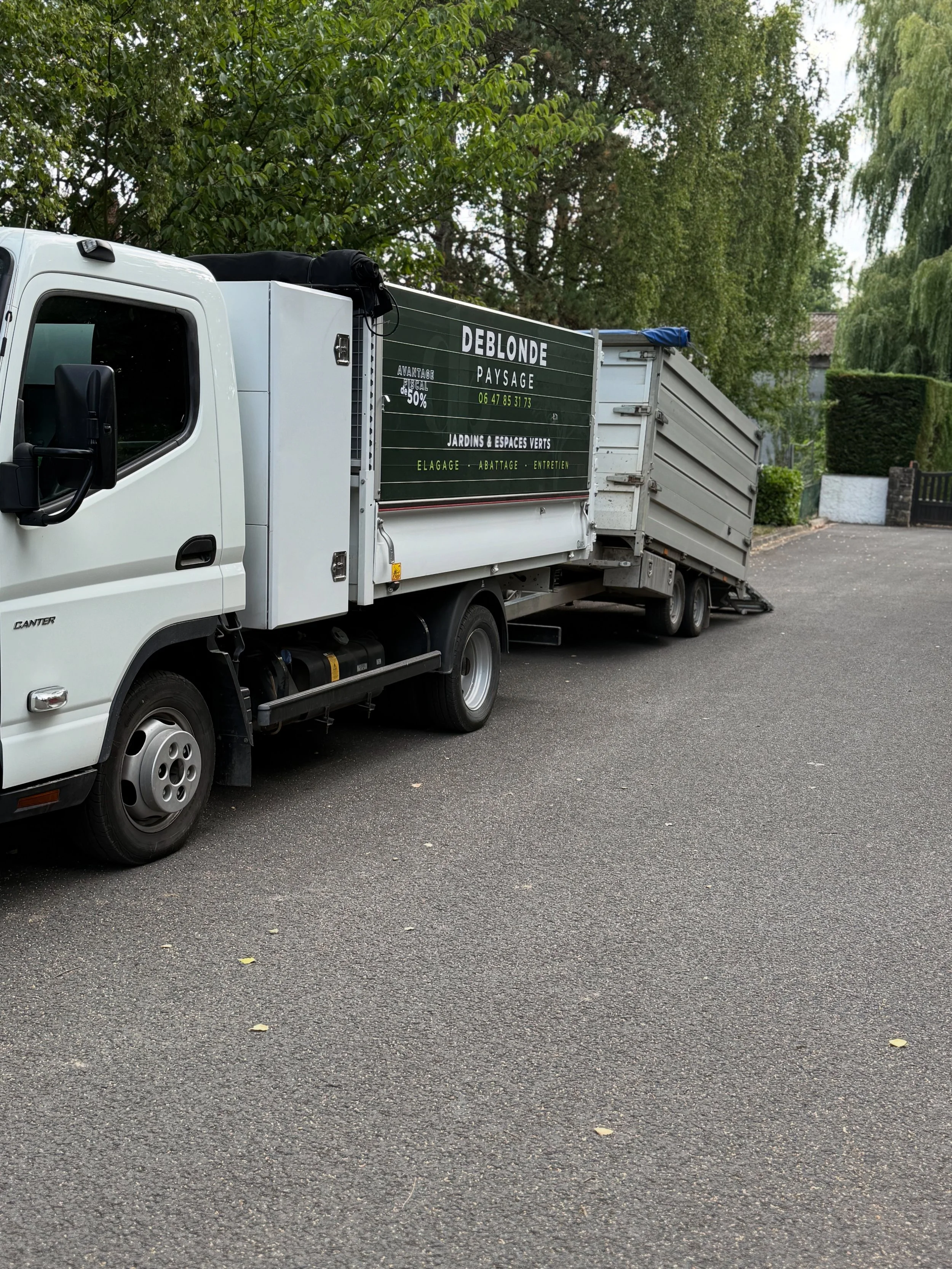 Camion blanc avec une remorque, stationné sur une route, entouré d'arbres et de haies, avec un panneau publicitaire sur la remorque pour une entreprise d'aménagement paysager nommée DEBLONDE, proposant des services de jardinage et d'entretien.