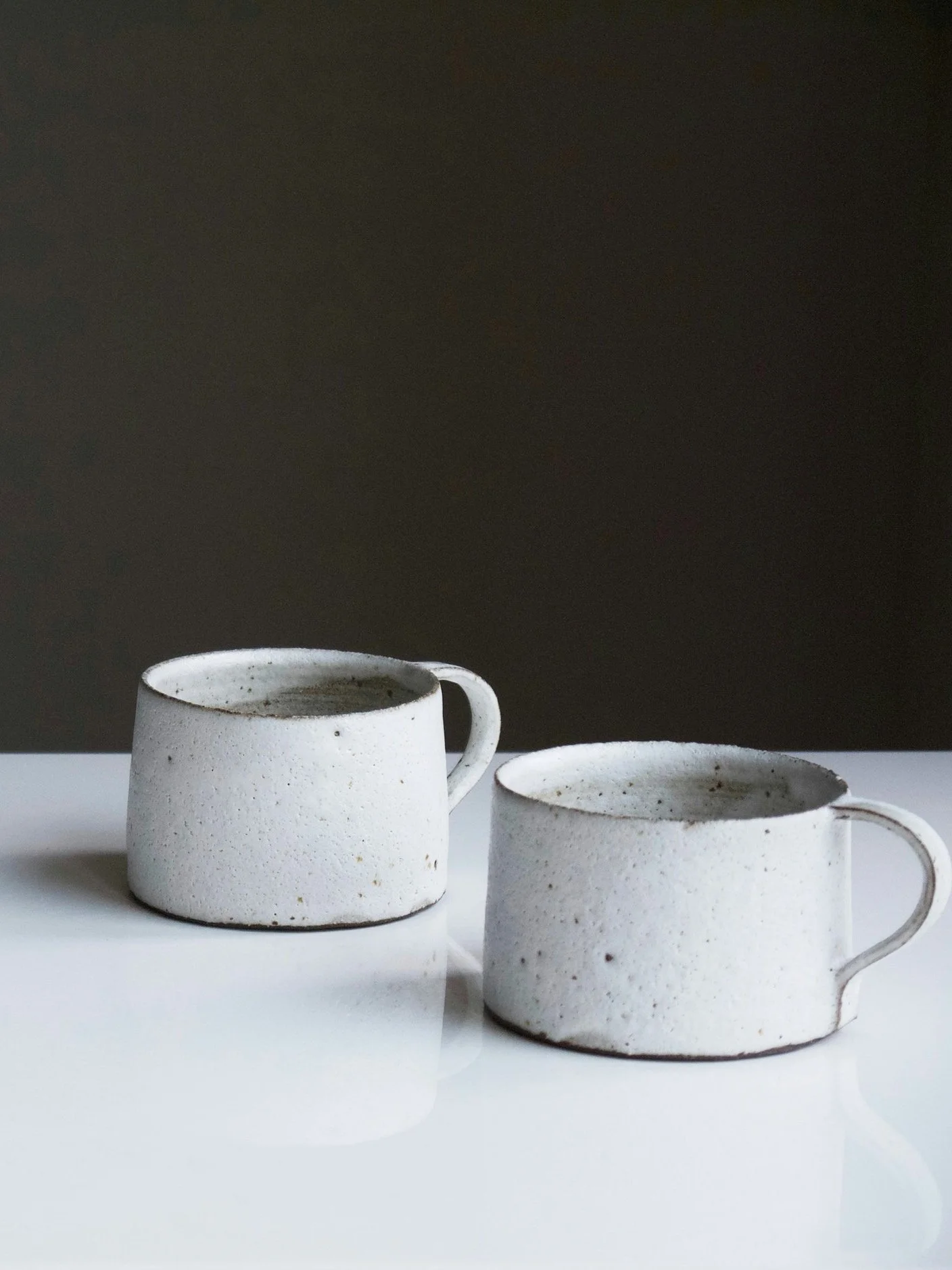 Two white ceramic coffee mugs, both filled with coffee and showing a rustic, speckled texture, placed on a white surface with a dark background.
