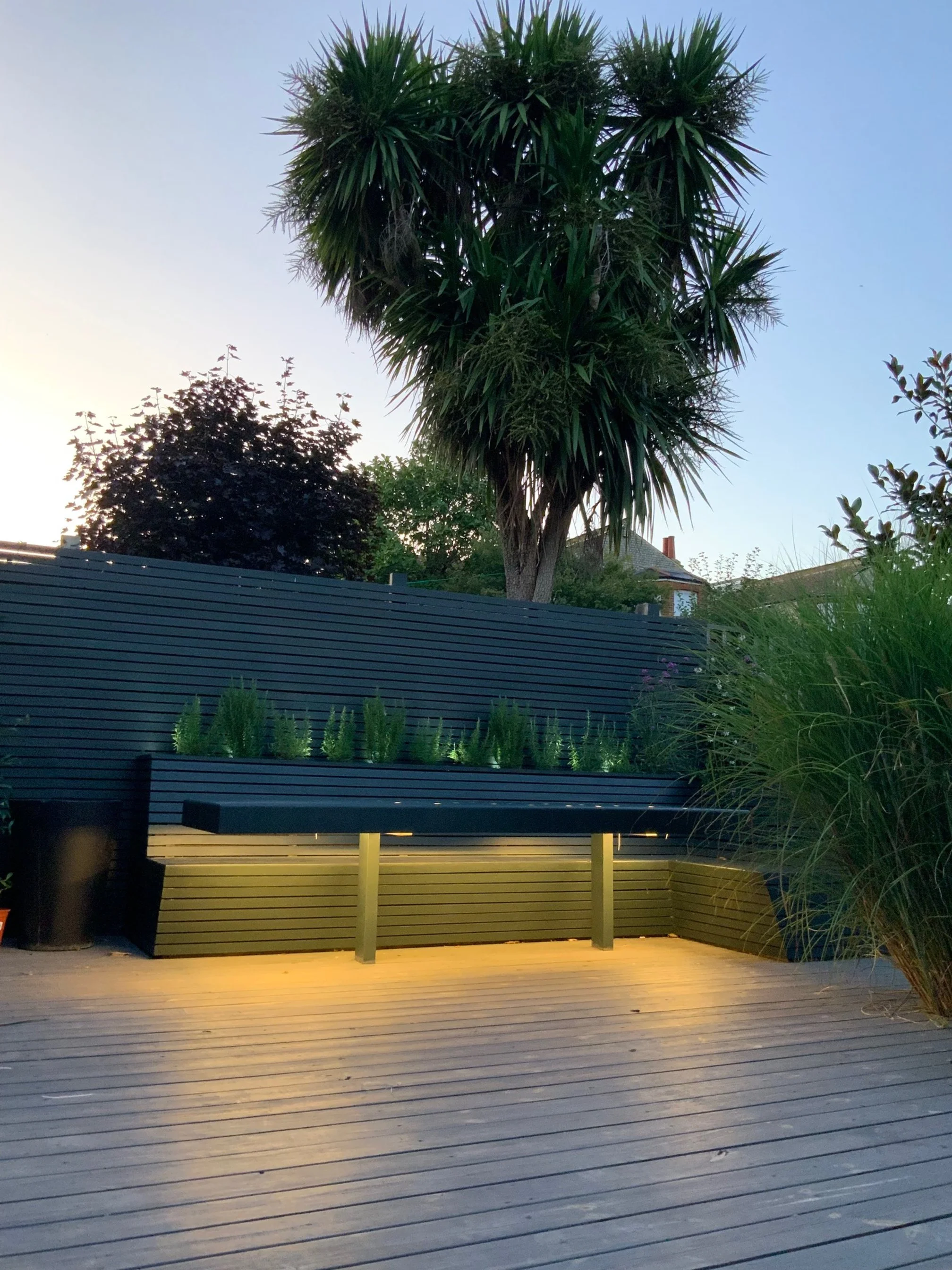 A modern outdoor wooden deck with built-in lighting, a long black bench with potted plants on top, a black fence, a tall palm tree, and various other trees and plants in the background during sunset.