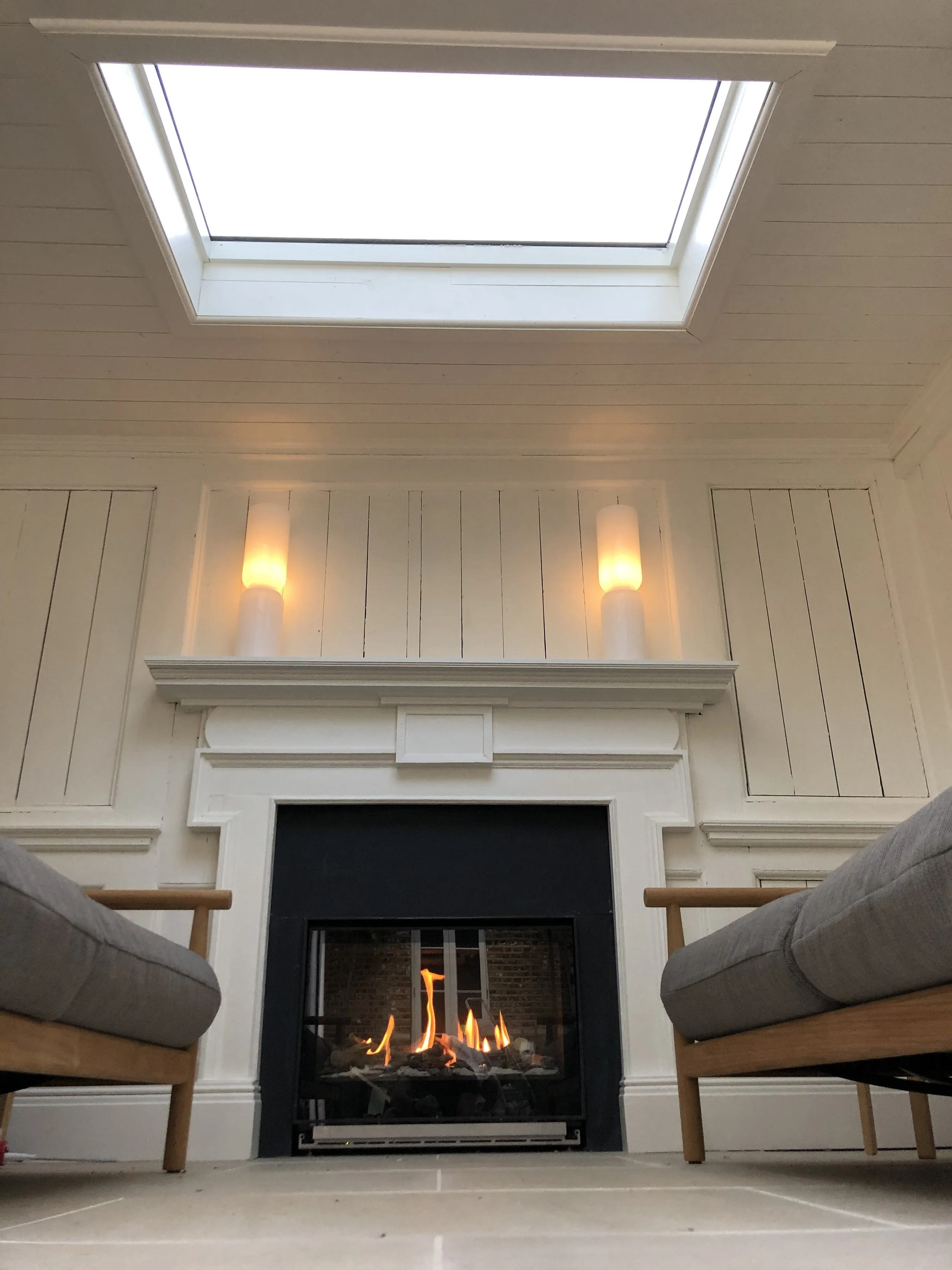 Living room with a fireplace, two chairs with gray cushions, wall sconces, a ceiling skylight, and a brick wall visible through the fireplace.
