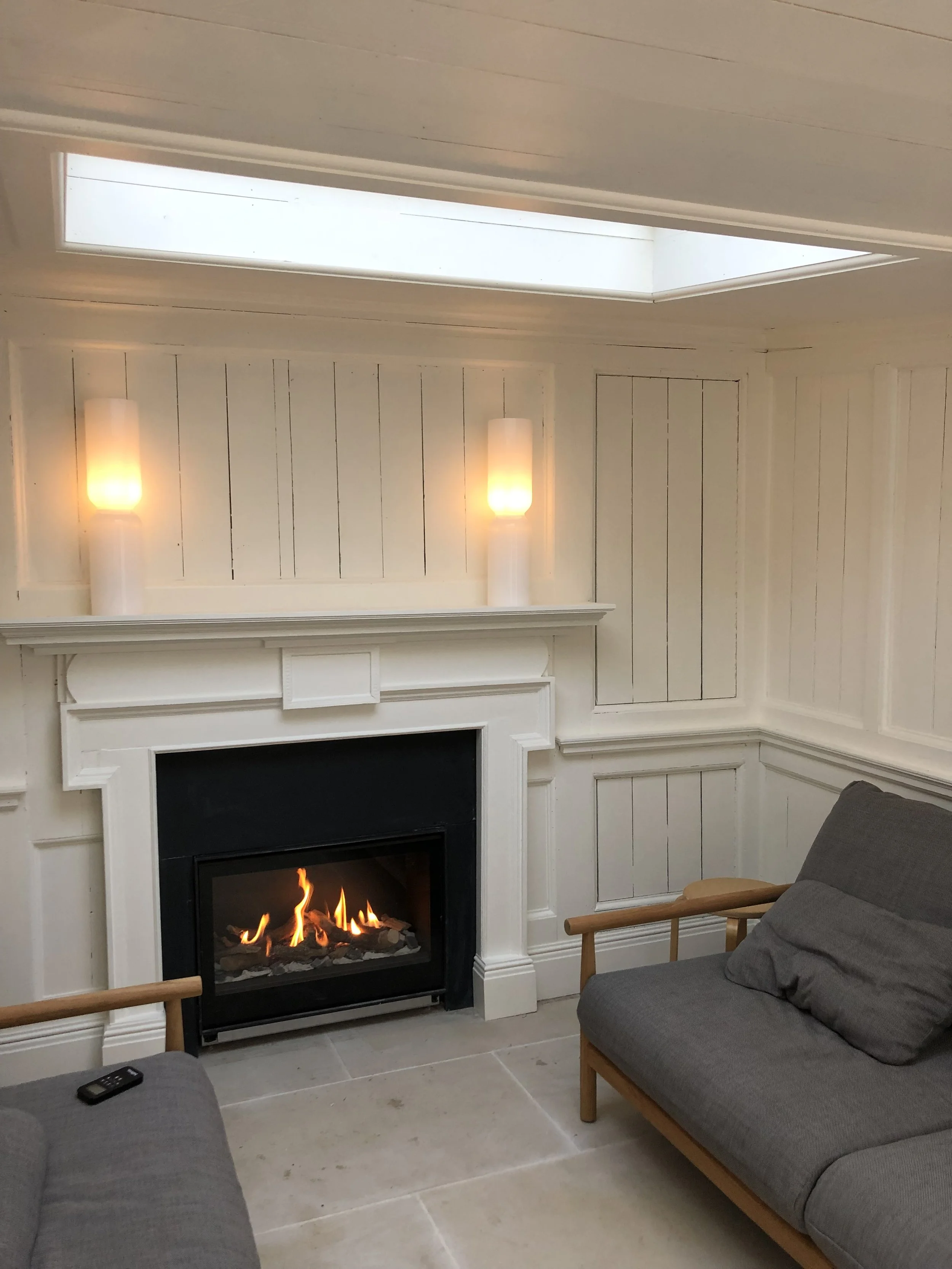 A cozy living room with a modern fireplace, gray couches, a remote control, wall-mounted lights, and a skylight.