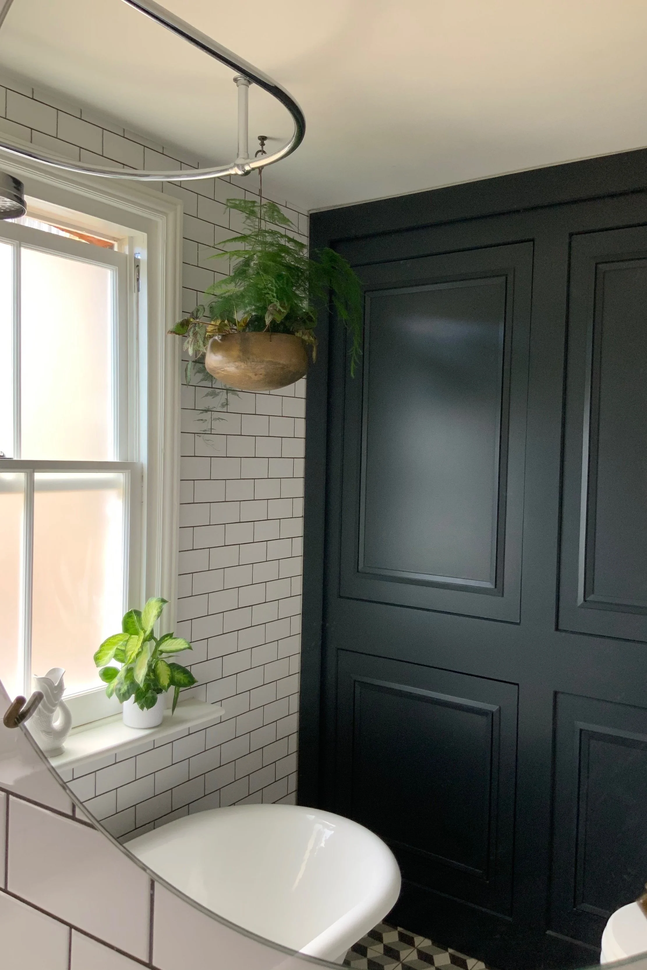 A bathroom with black cabinets, white subway tile walls, and a window with a plant on the sill. A hanging potted plant is above, and a clawfoot tub is partially visible.