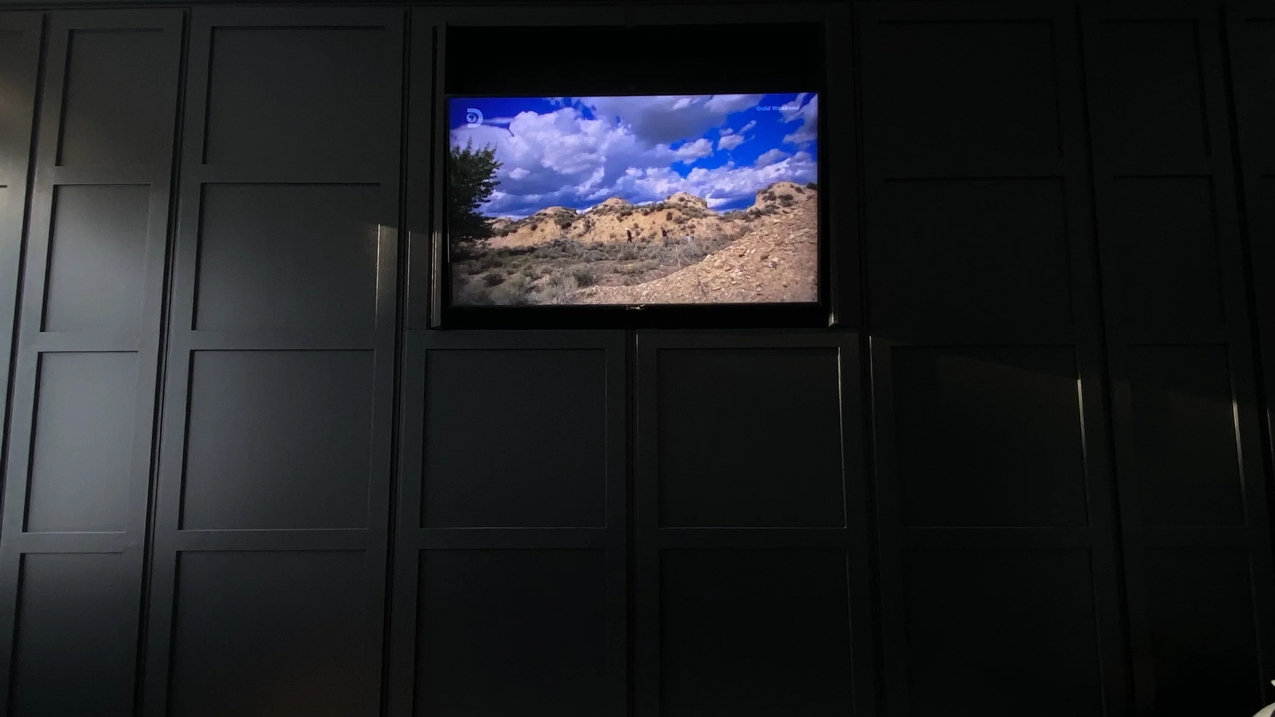 The image shows a television mounted on a black wall, displaying a landscape with a rocky terrain, shrubs, and blue sky with clouds.