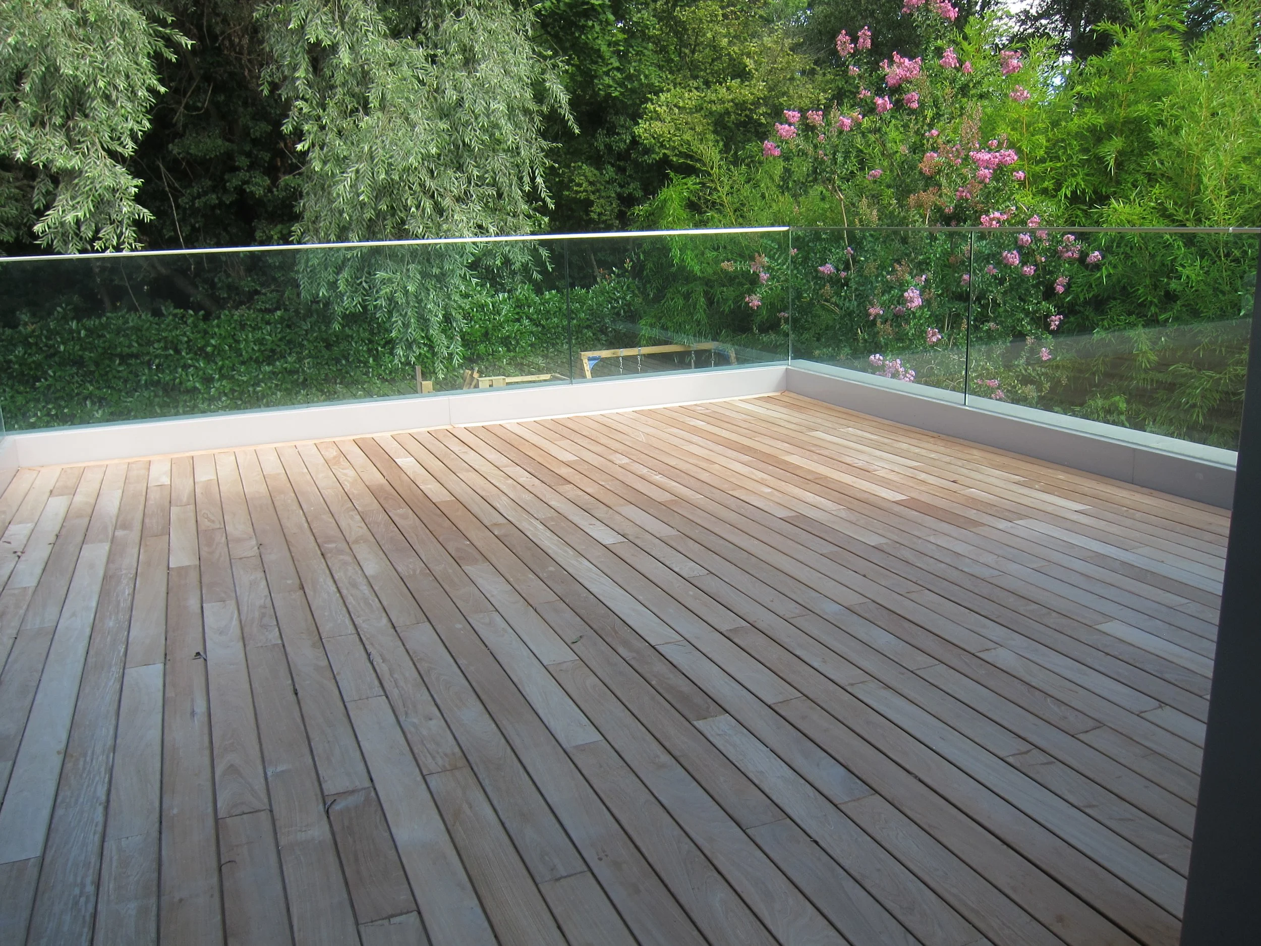 Empty wooden deck with glass railing overlooking green trees and pink flowers.