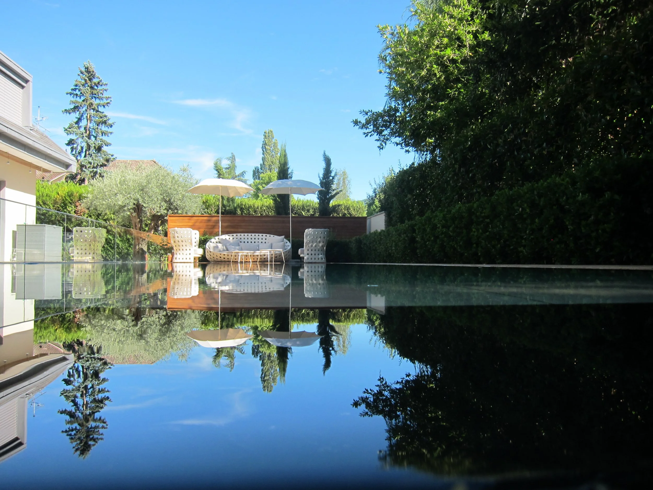 Outdoor patio with a pool, white lounging chairs, and umbrellas under a blue sky, surrounded by trees and bushes