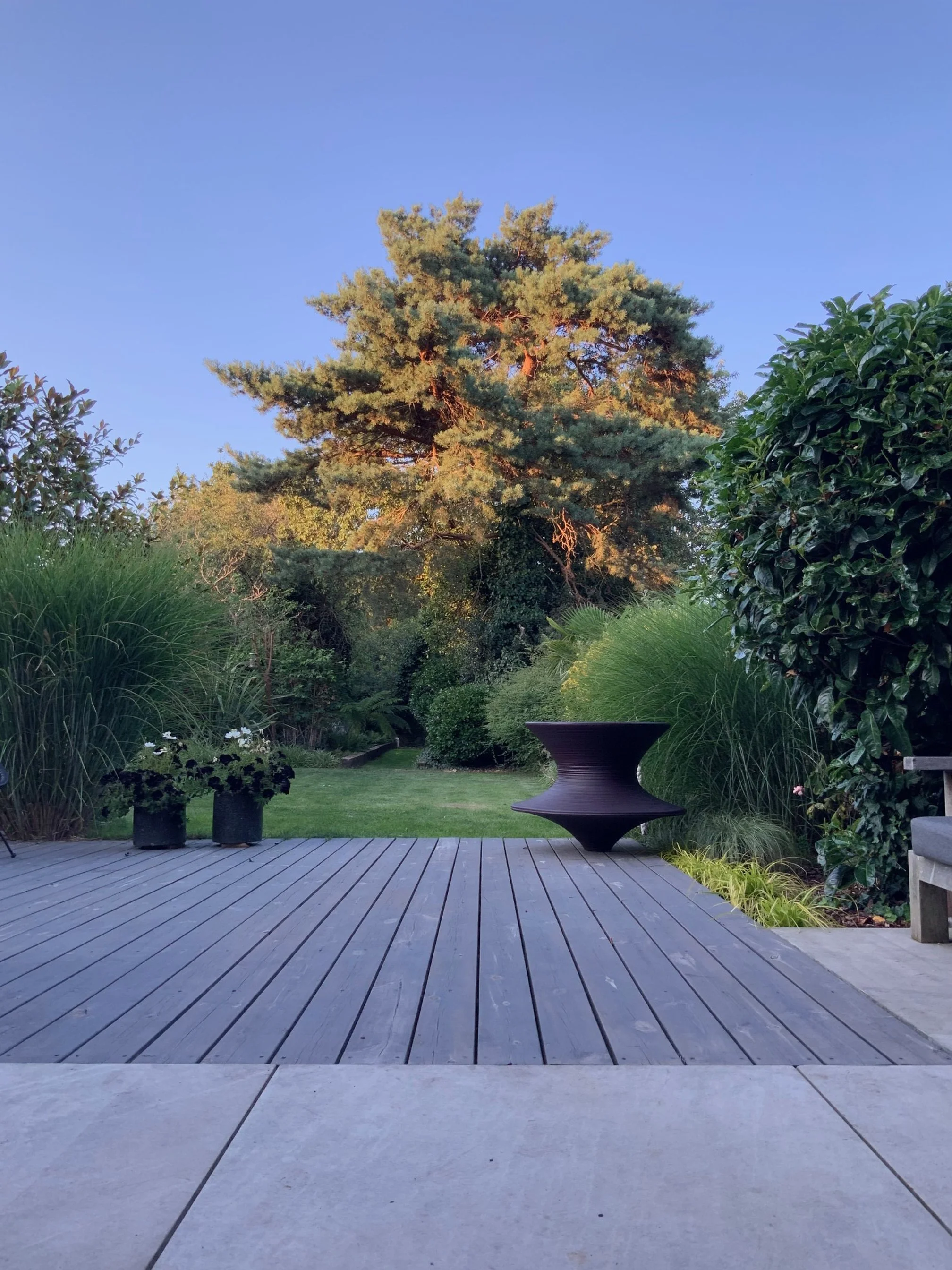 A wooden deck with potted plants and a modern black outdoor table in a lush, green backyard with tall trees and shrubs under a clear blue sky.