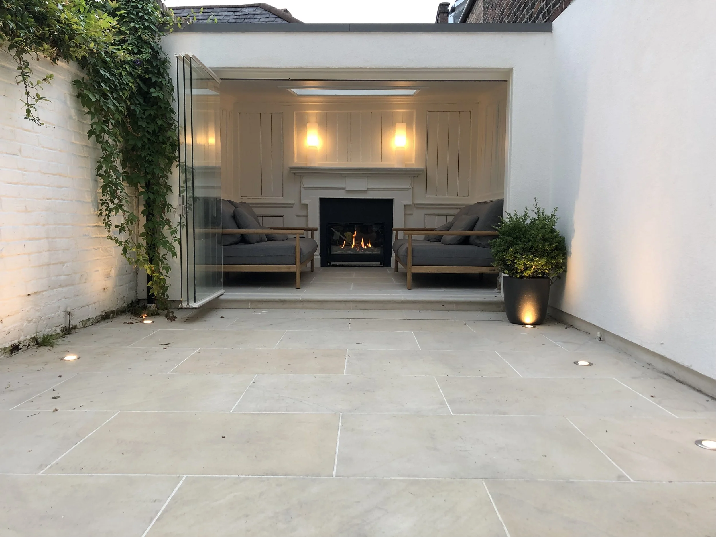 Outdoor patio with beige stone tiles, white brick and white wall, led floor lights, potted plant with spotlight, and a cozy seating area with a fireplace, two grey couches, and lighting fixtures.