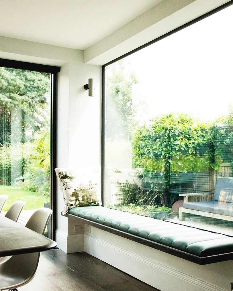 Bright corner of a modern dining room with large floor-to-ceiling glass windows, a cushioned window seat with a floral throw pillow, and views of a lush backyard with trees and outdoor furniture.