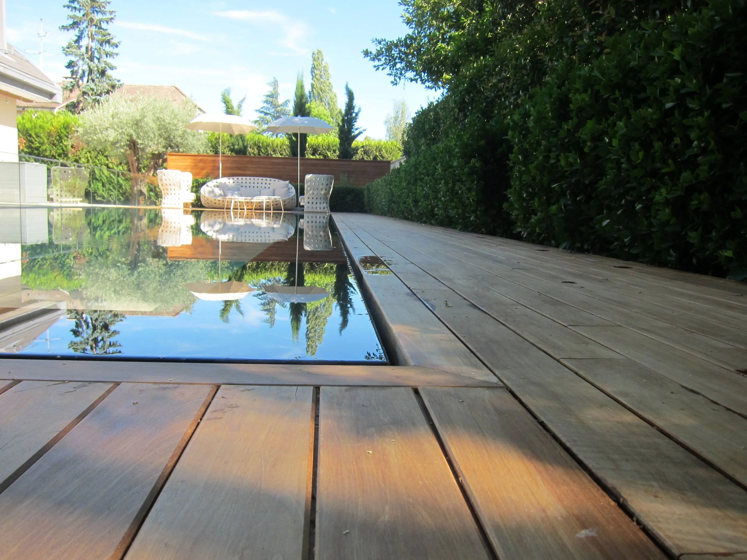 Backyard pool with wooden deck, surrounded by green bushes and outdoor furniture including two umbrellas, a lounge chair, and a curved seating area, with trees and houses in the background under a blue sky.