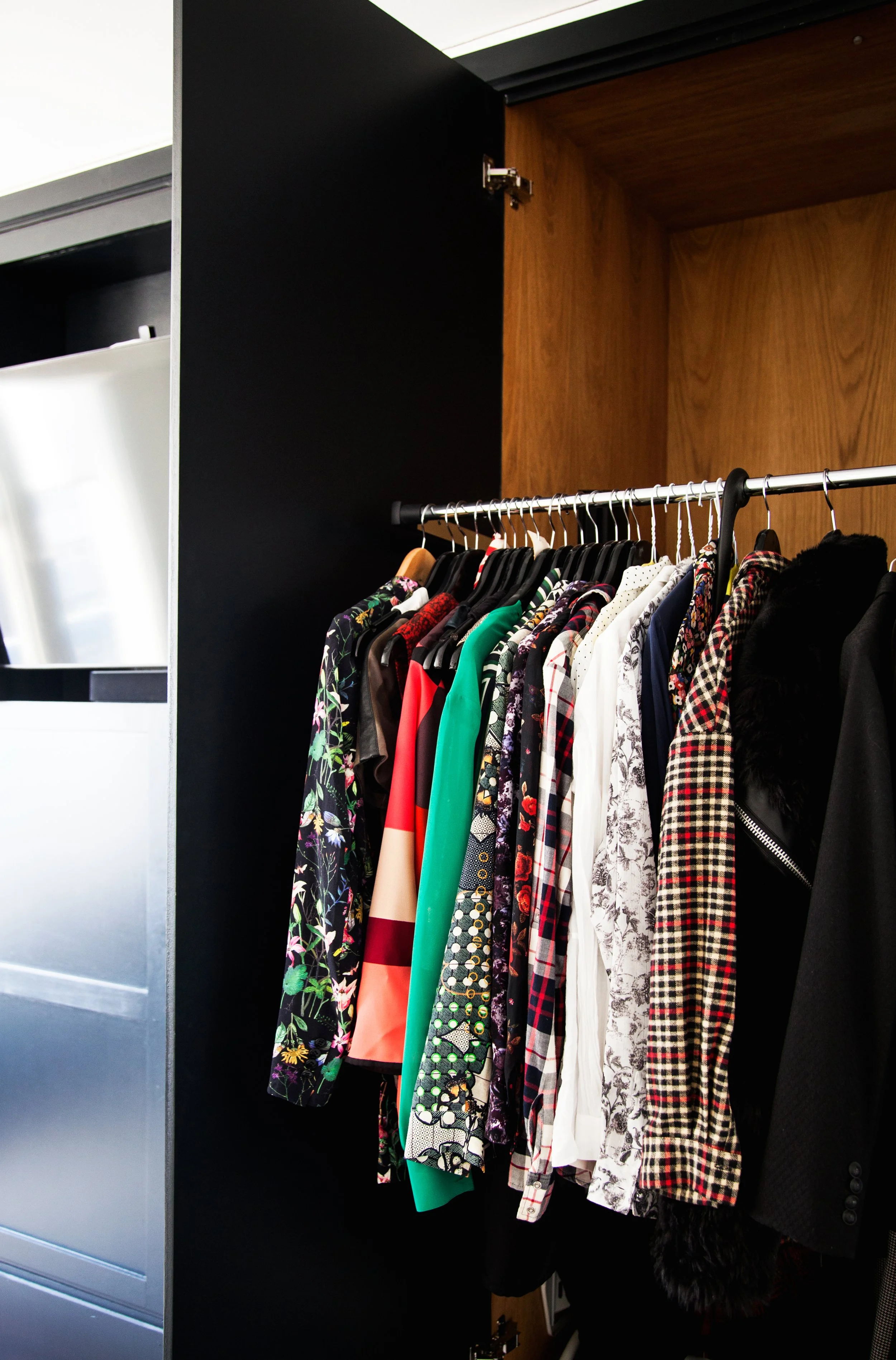 A closet with a black door open, revealing a clothing rack with various colorful and patterned shirts and jackets hanging on hangers.