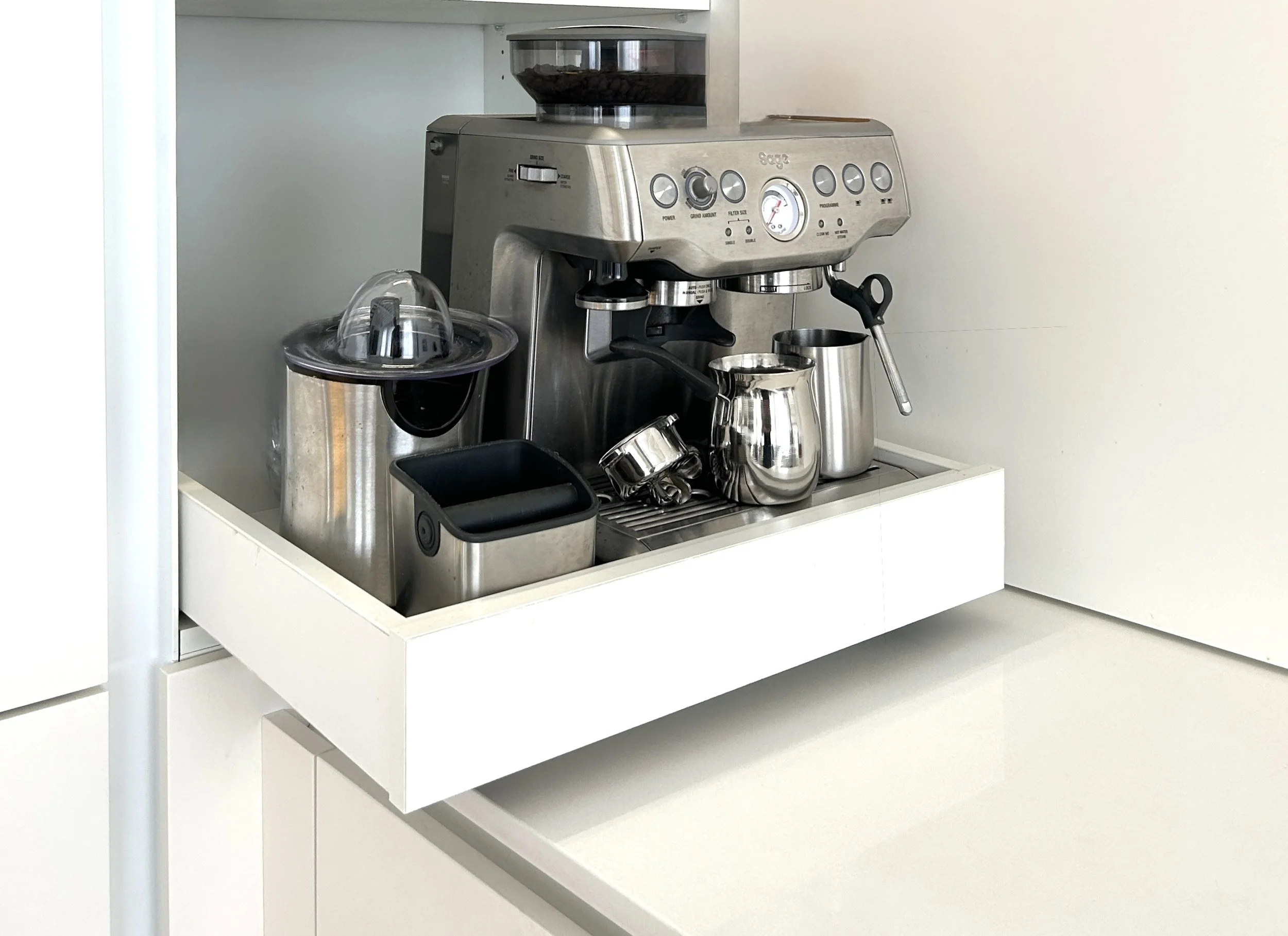 A silver espresso machine on a white kitchen counter with various stainless steel coffee accessories around it.