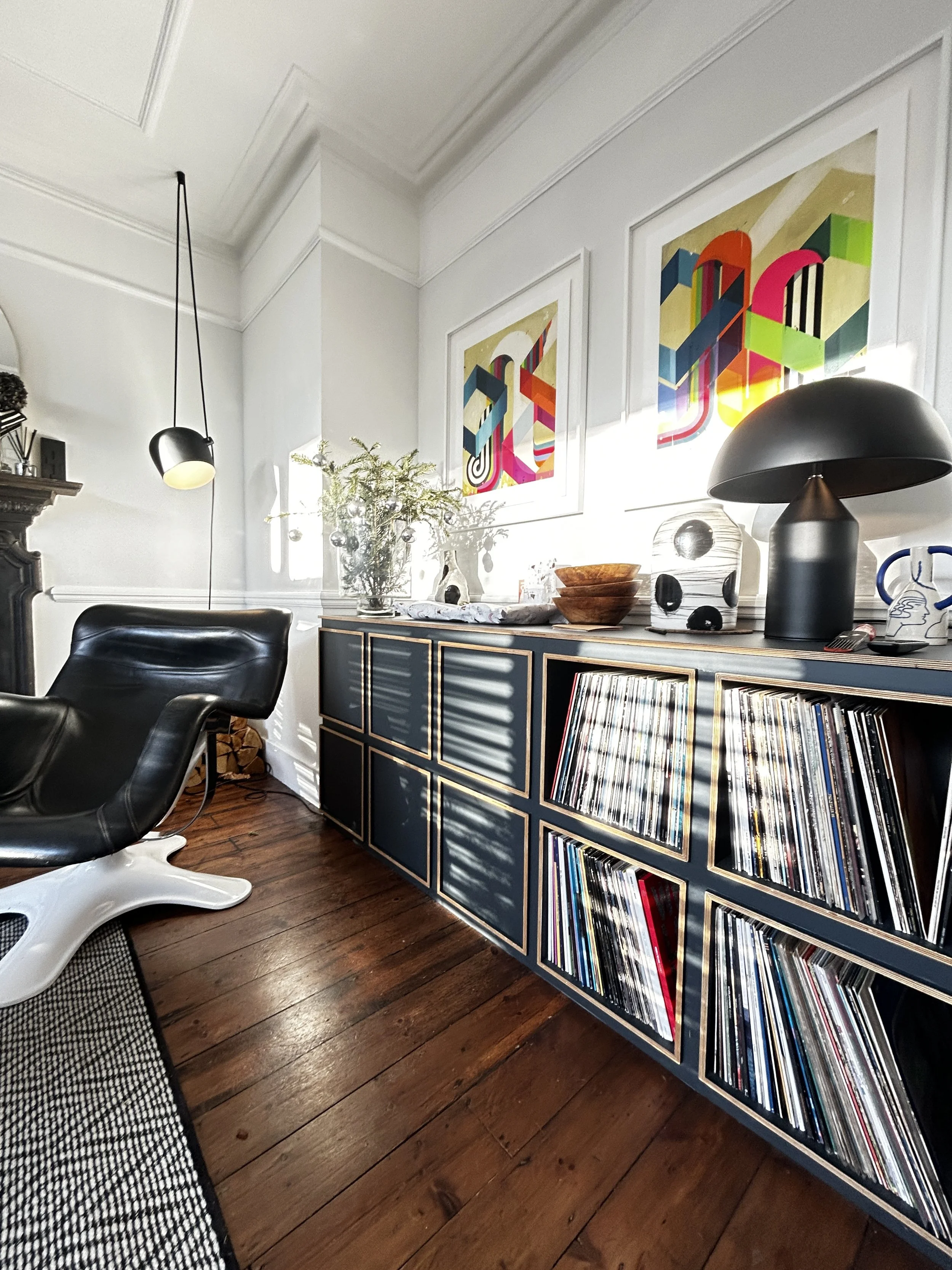 A living room corner with a black leather lounge chair, a blue mid-century modern cabinet with gold trim, and wall art with colorful geometric designs. There are framed abstract artworks, a table lamp, and various decorative items on top of the cabinet, with sunlight casting shadows.