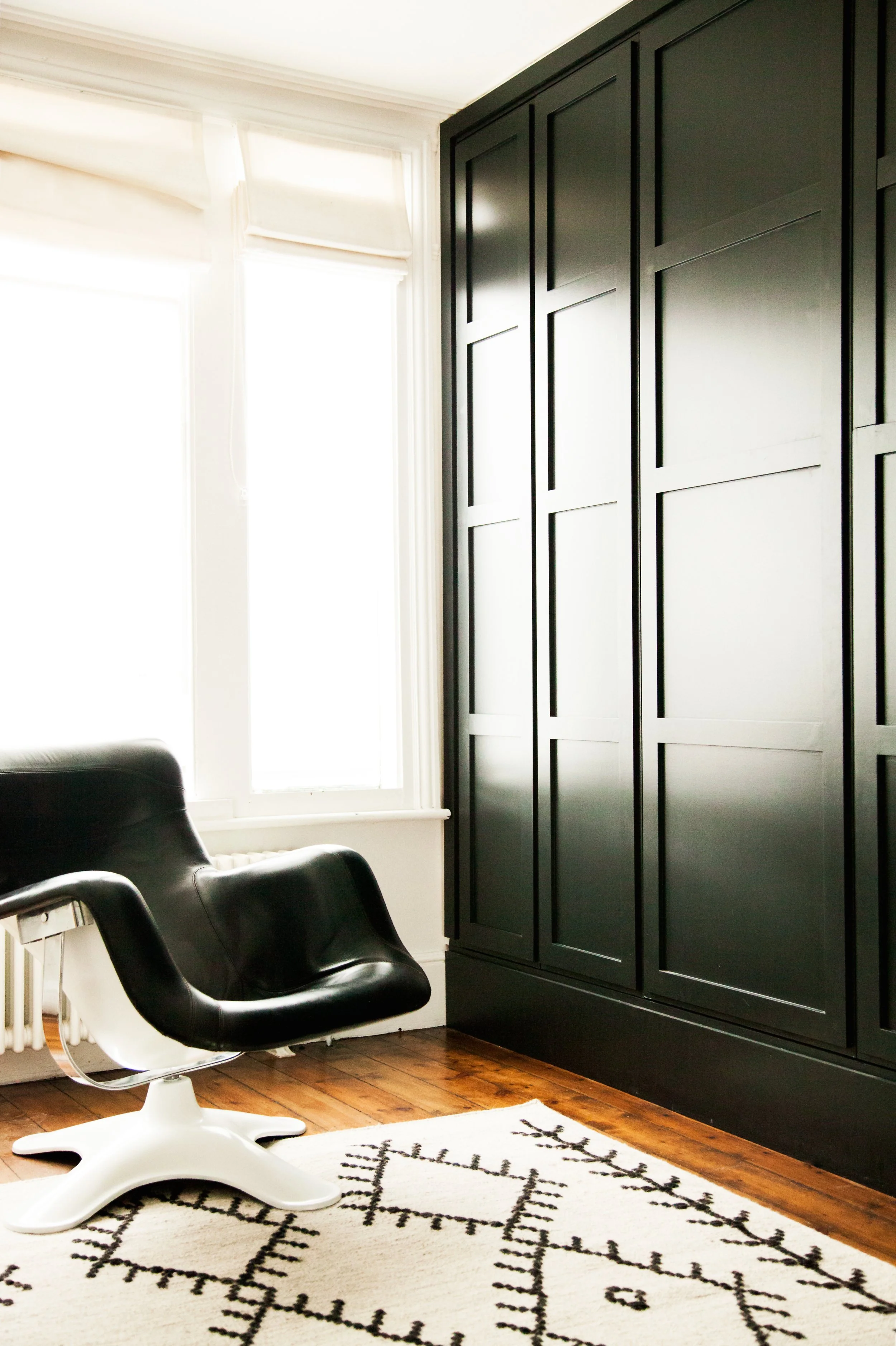 A black leather lounge chair next to a large black wardrobe in a room with wooden flooring, a white rug, and two large windows with white blinds.