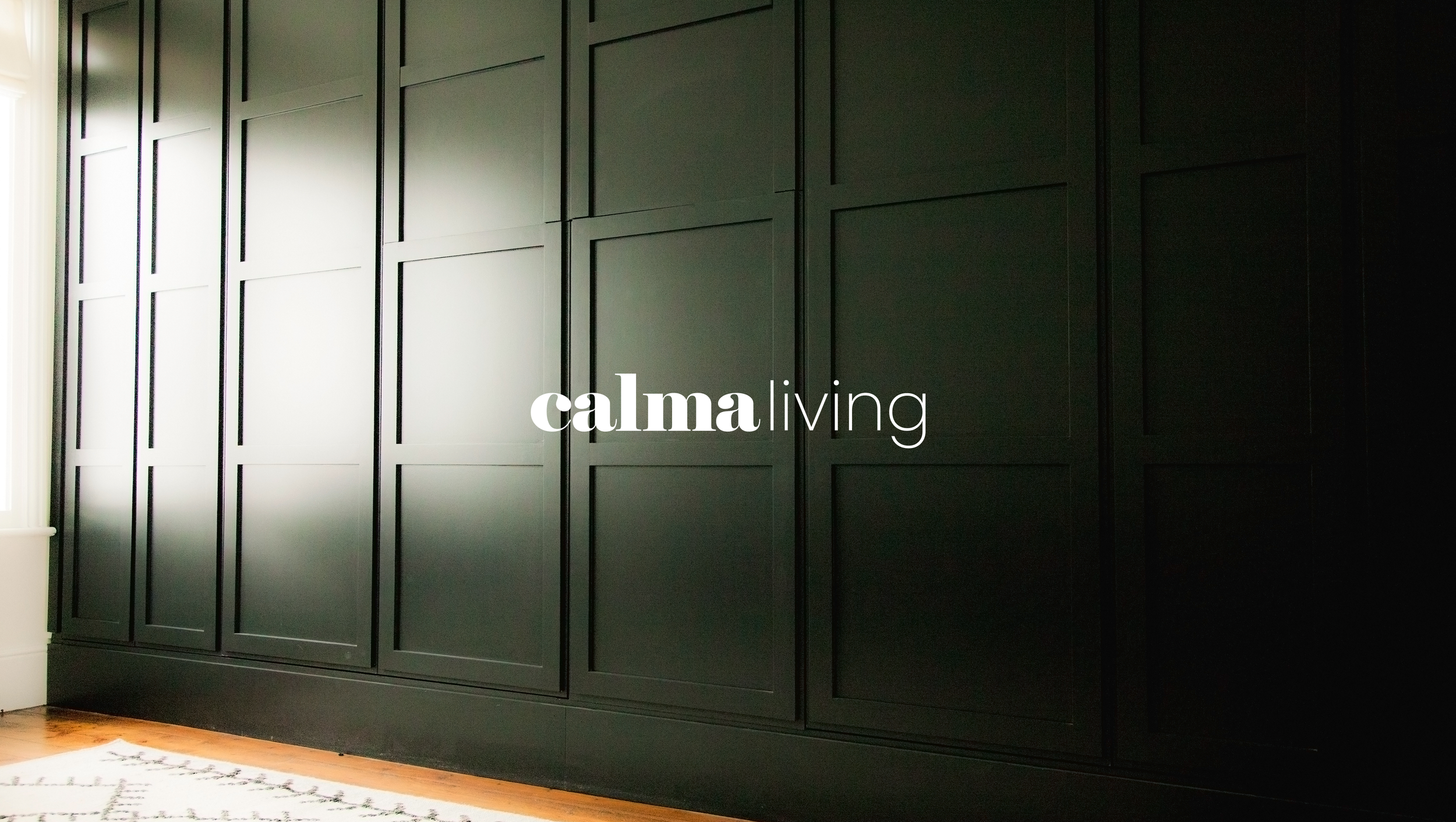 Dark green paneled wall with a white curtain and a portion of a white rug with a black geometric pattern on a wooden floor.