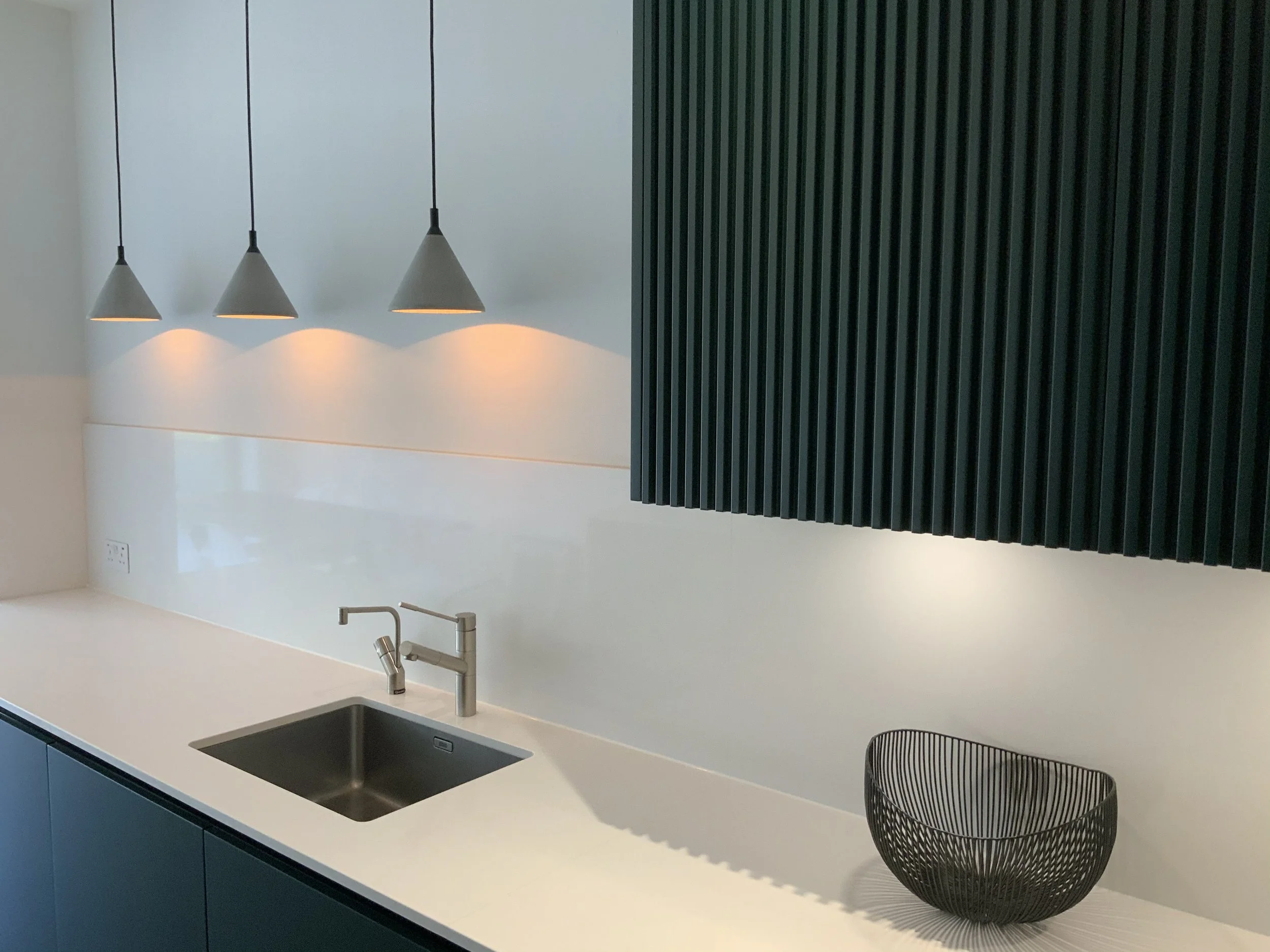 Modern kitchen with three cone-shaped pendant lights, a white countertop, a stainless steel sink and faucet, dark green cabinets, and a black wire fruit basket.