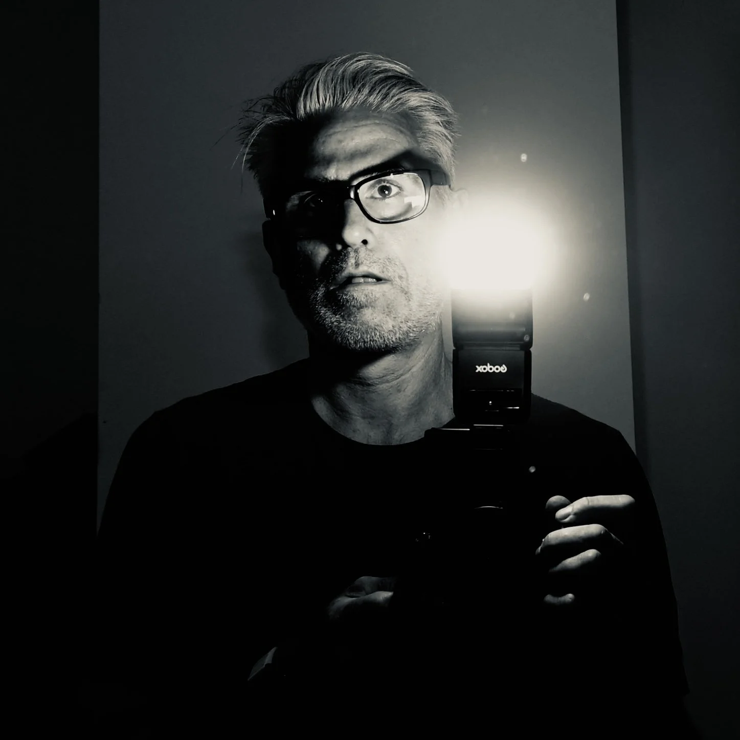 Black and white photo of a man with glasses taking a selfie in a mirror, with a bright flash illuminating part of his face.