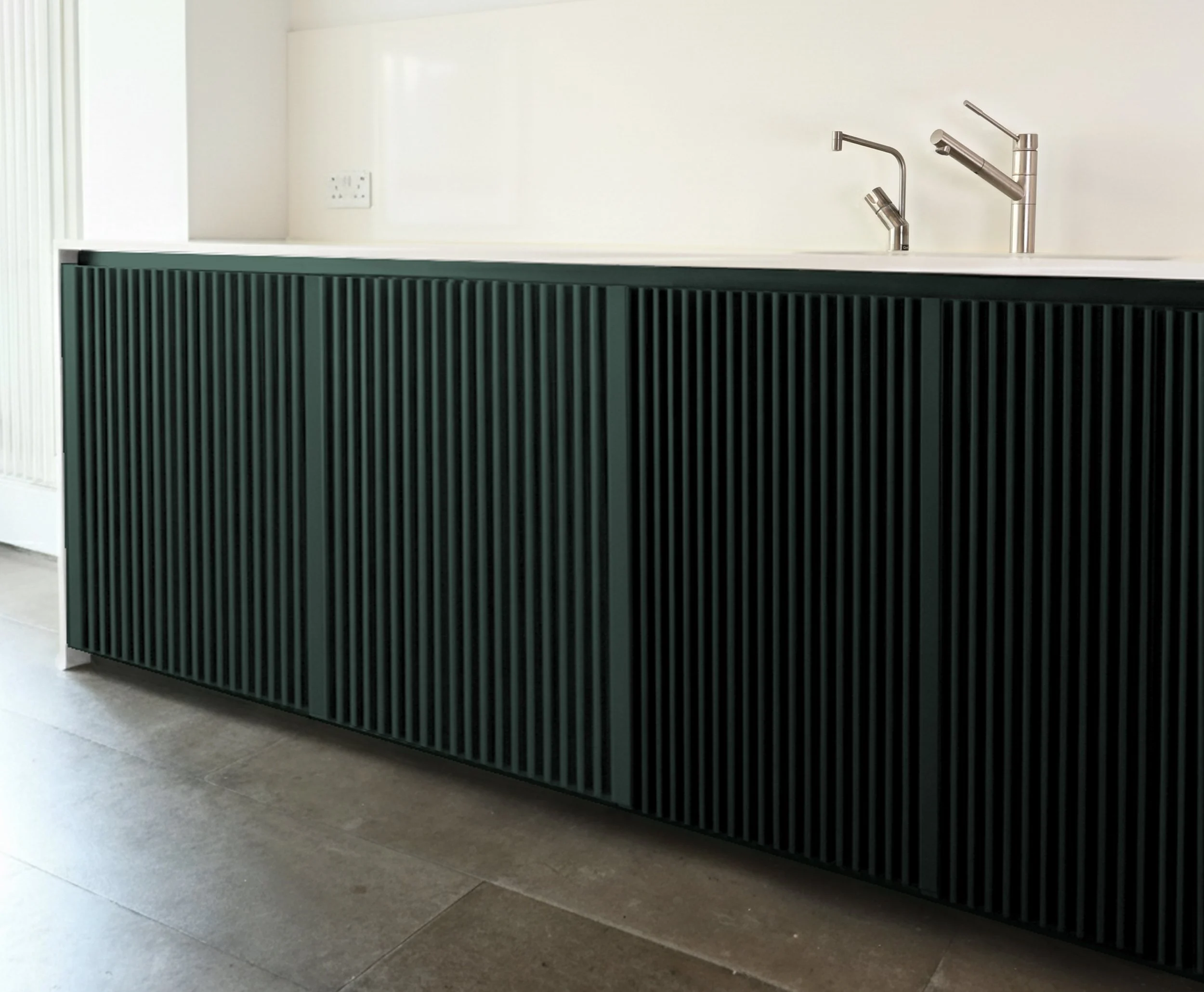 Modern kitchen island with a green slatted front panel and stainless steel sink fixtures against a white wall with an electrical outlet.