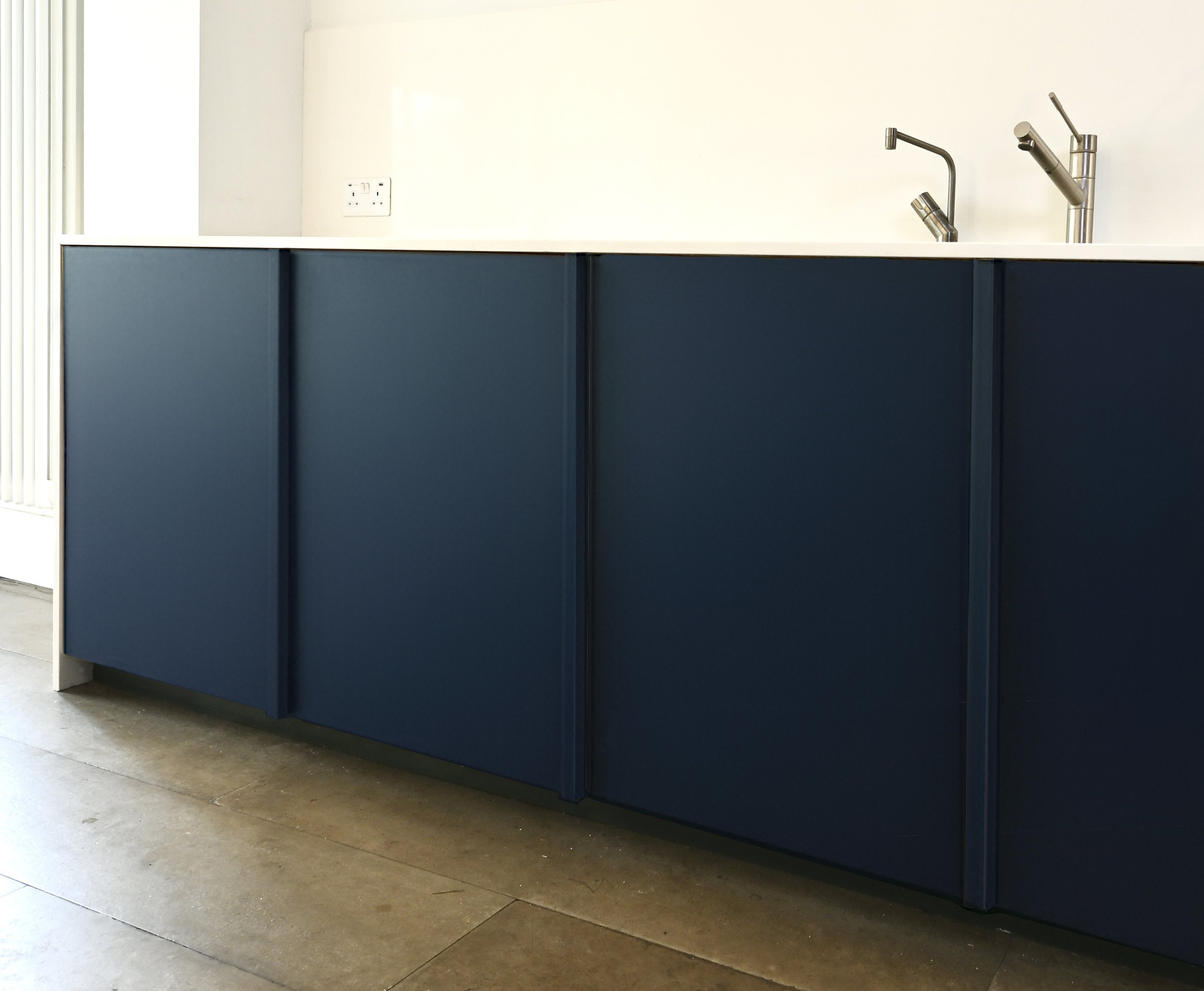 Modern kitchen cabinet with blue doors and a white countertop, featuring a double kitchen sink with two faucets and a white wall with an electrical outlet.