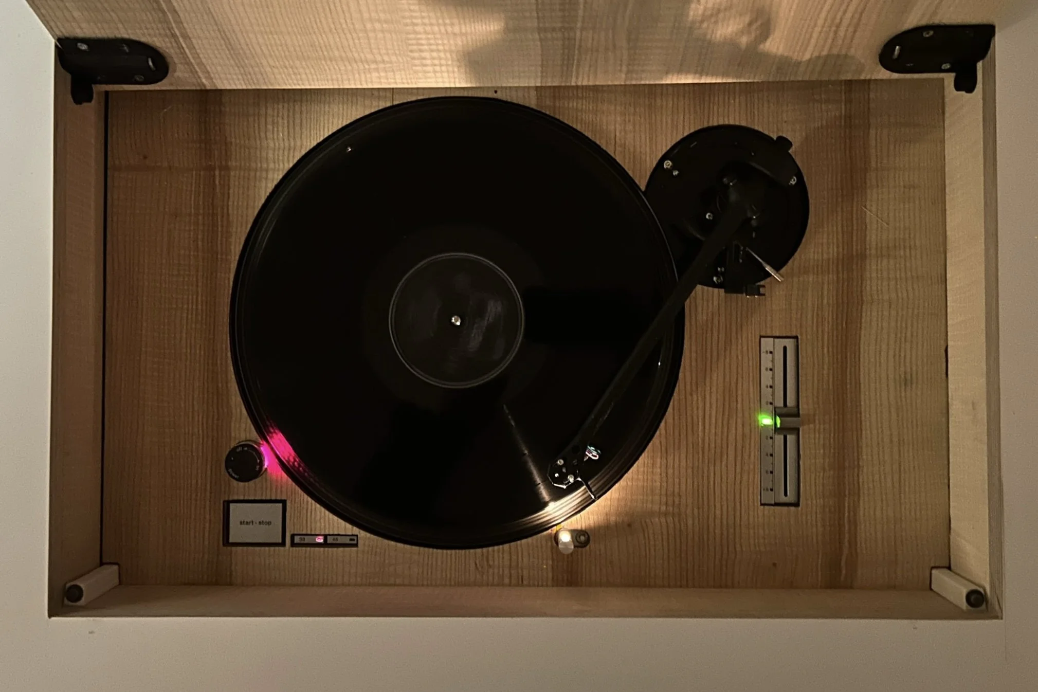 Top view of a vintage turntable with a wooden base, black platter, tonearm, and control buttons, placed inside a wooden enclosure.