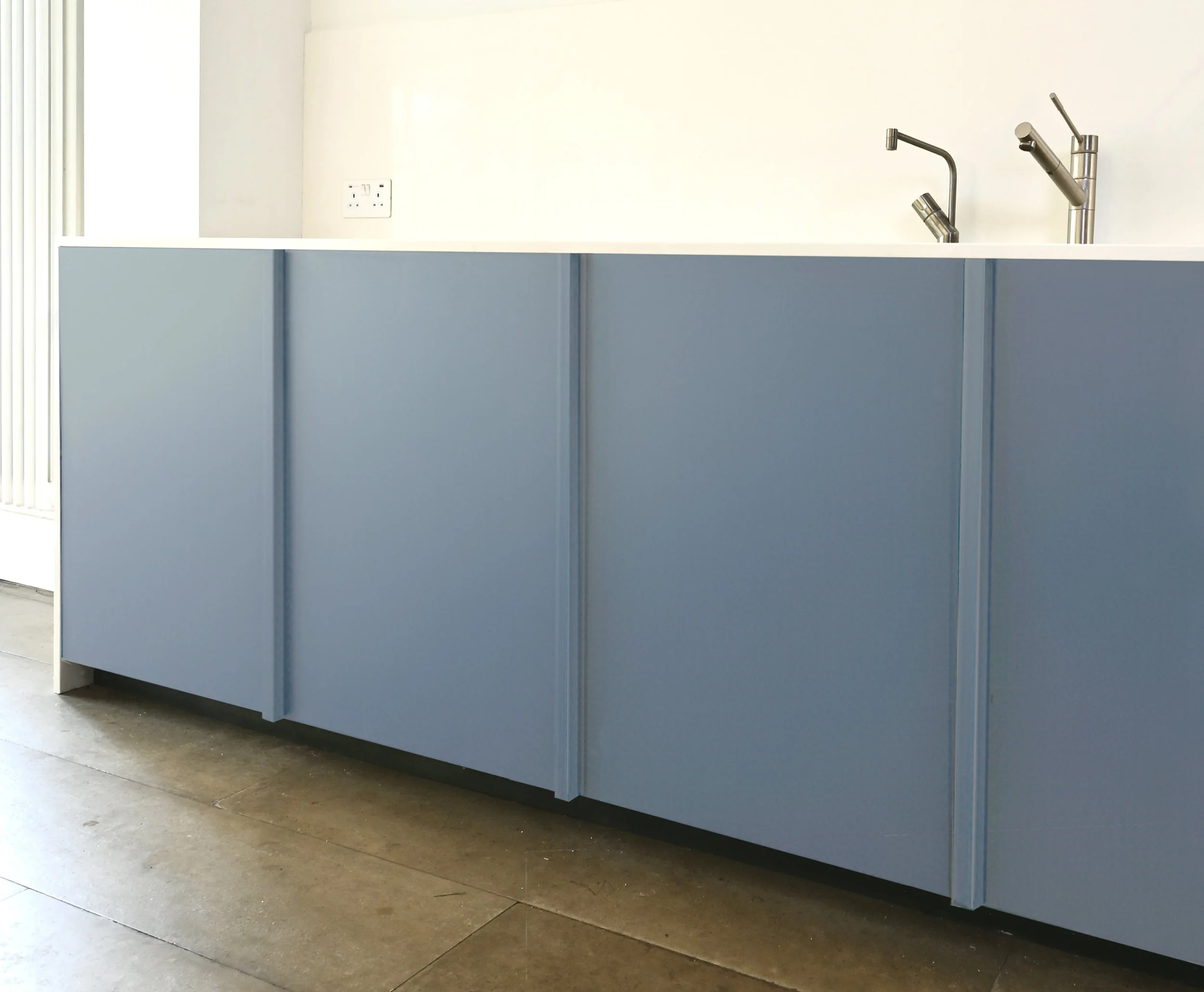 Blue kitchen cabinet with a white countertop and a stainless steel faucet and sink, with a white wall and electrical outlet in the background.