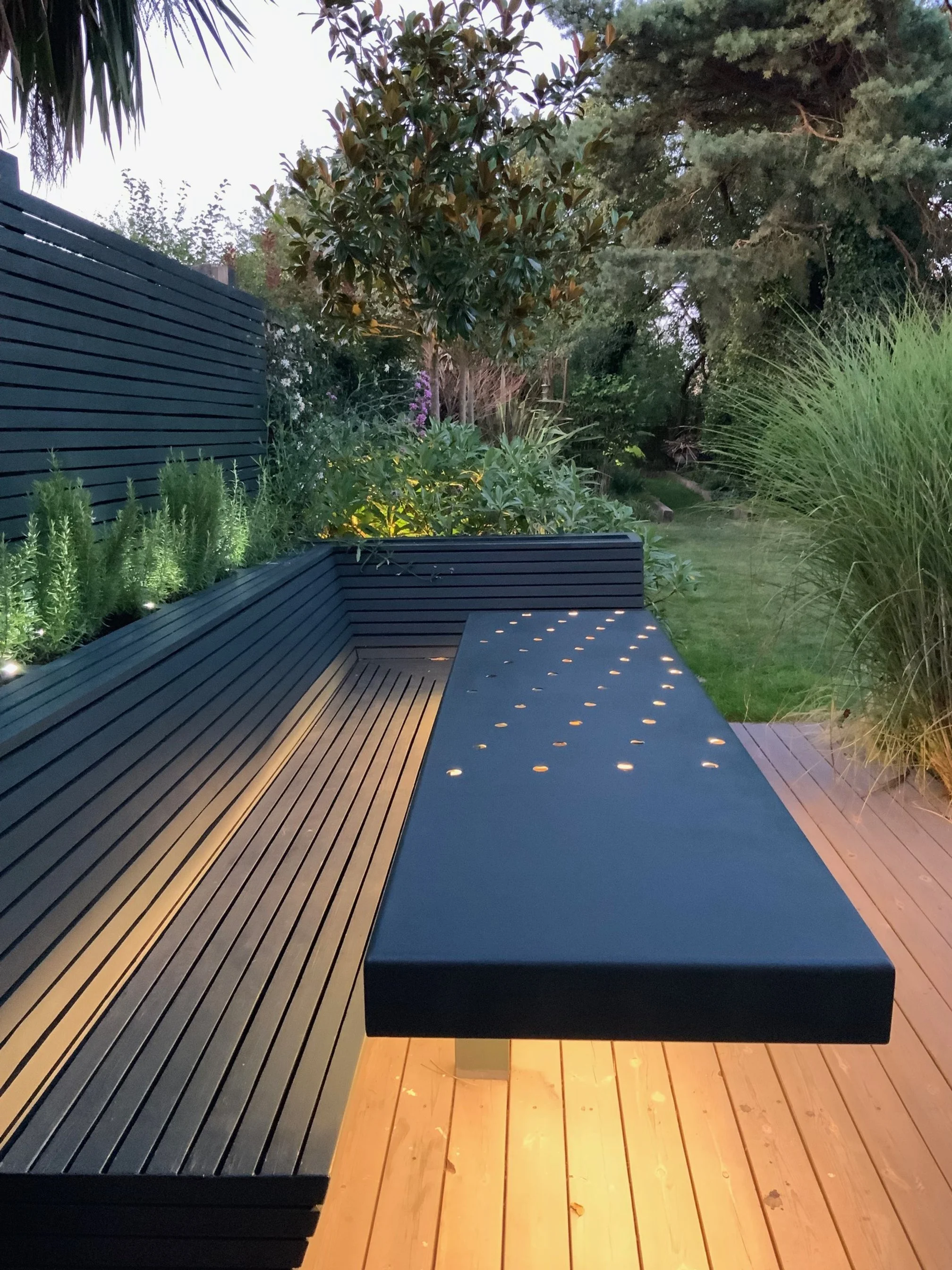 Outdoor patio with modern black metal bench with built-in lighting, surrounded by lush greenery, trees, and garden plants at dusk.
