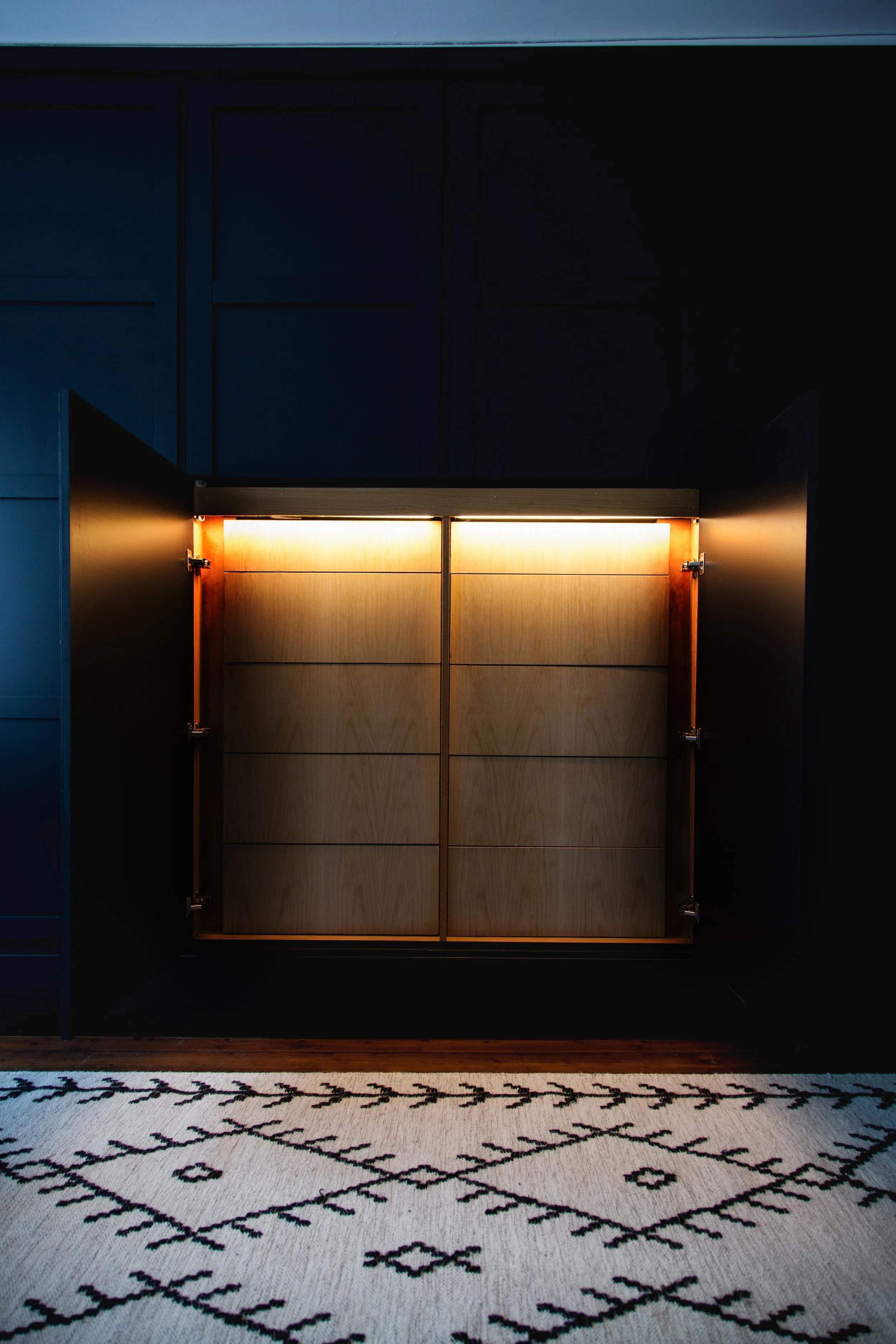 Open cabinet doors revealing illuminated wooden interior in a dark room with patterned rug.