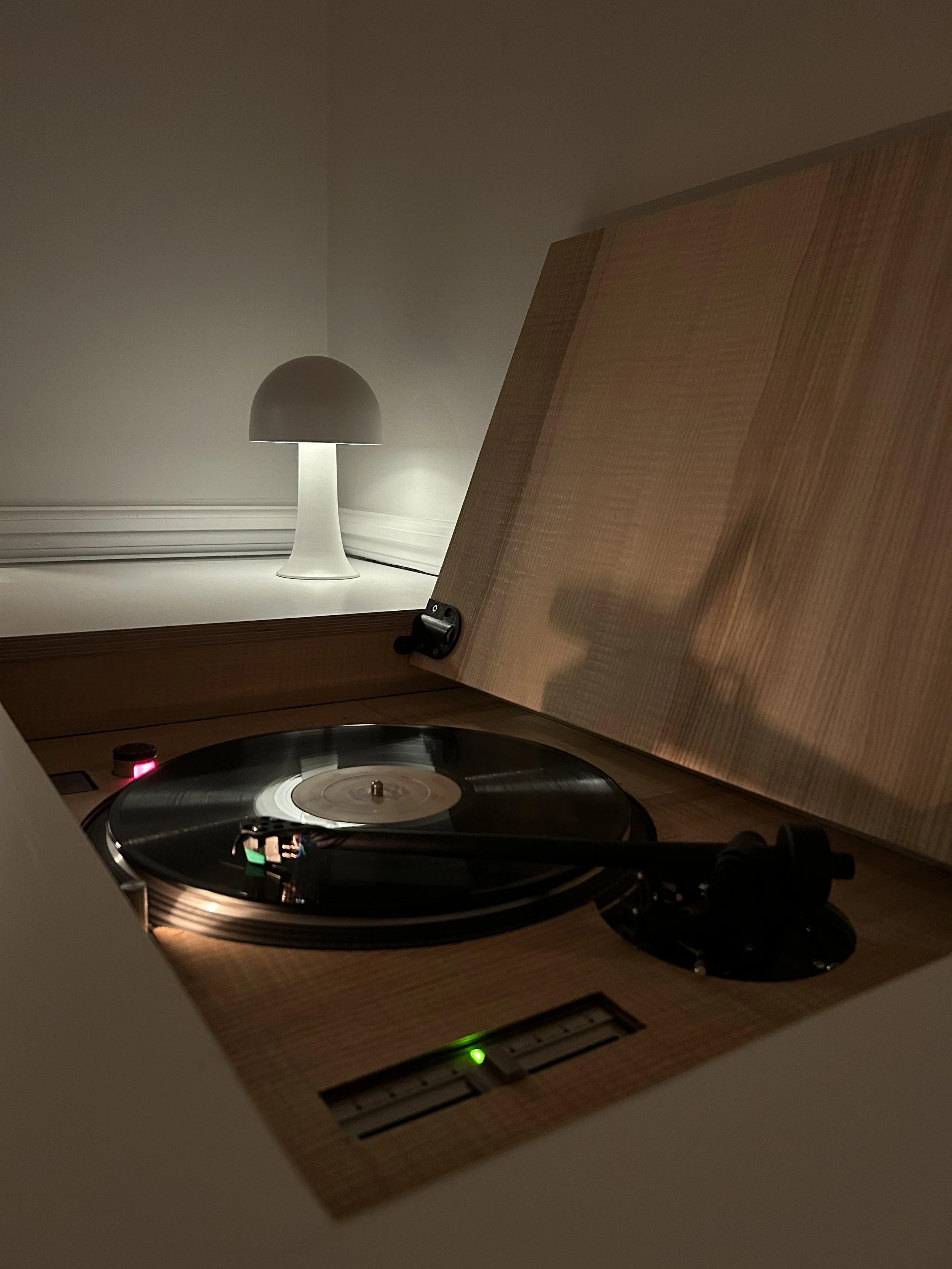 A record player with a spinning vinyl on a wooden turntable, partially opened lid, illuminated by a small grade light, with a modern white lamp in the background on a shelf.