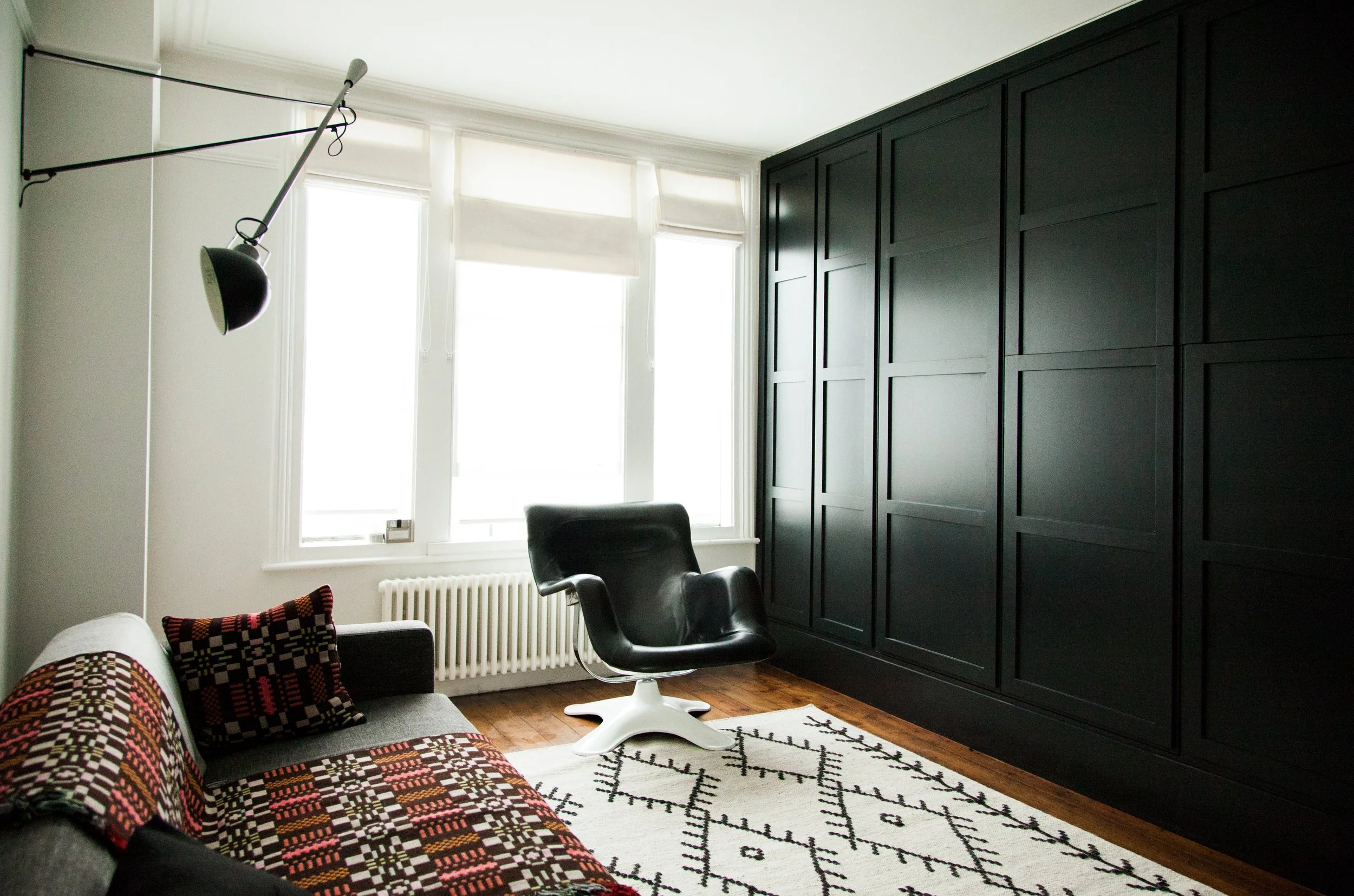 A brightly lit room with large windows, a black wardrobe, a black modern chair, a gray sofa with colorful patterned pillows, a white rug with black geometric designs, and a ceiling-mounted lamp or light fixture.