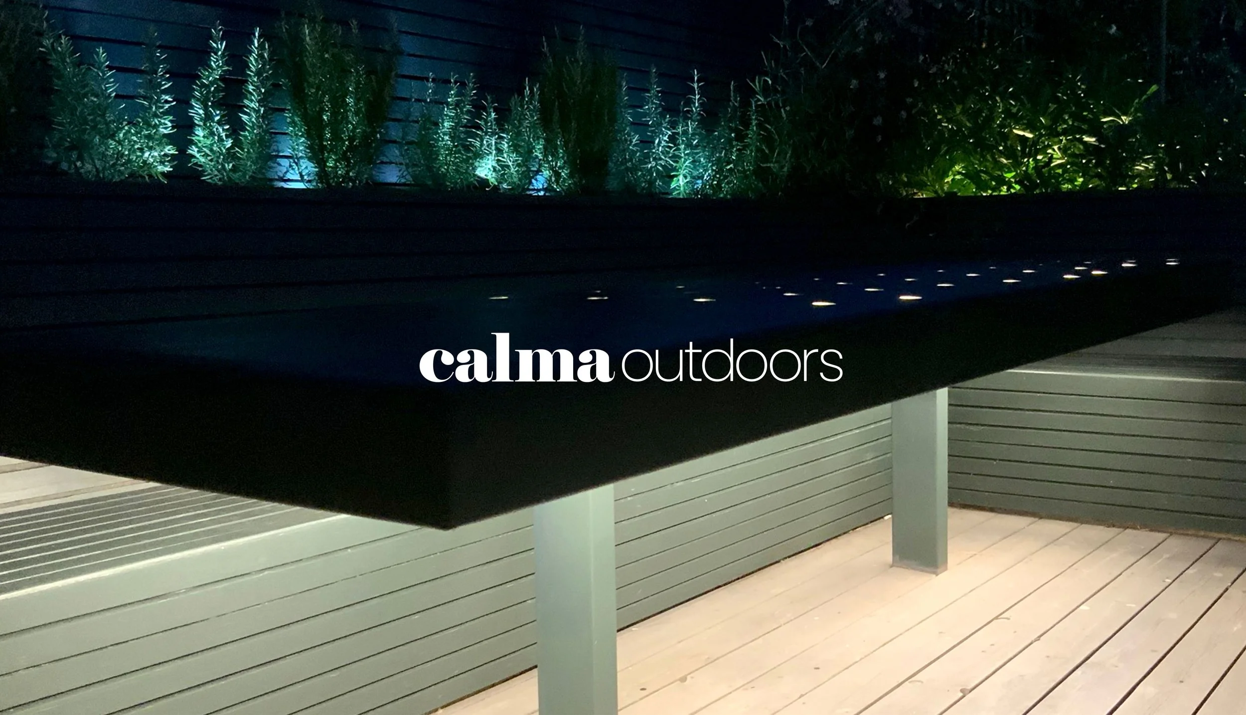 Outdoor deck with a black table and green plants illuminated by outdoor lighting at night, with the text 'calma outdoors' overlaid.