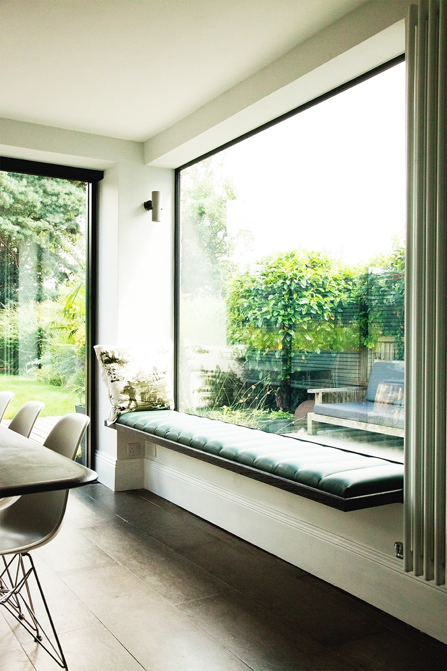A bright indoor room with large floor-to-ceiling windows overlooking a green outdoor garden, featuring a cushioned window seat and indoor modern decor.