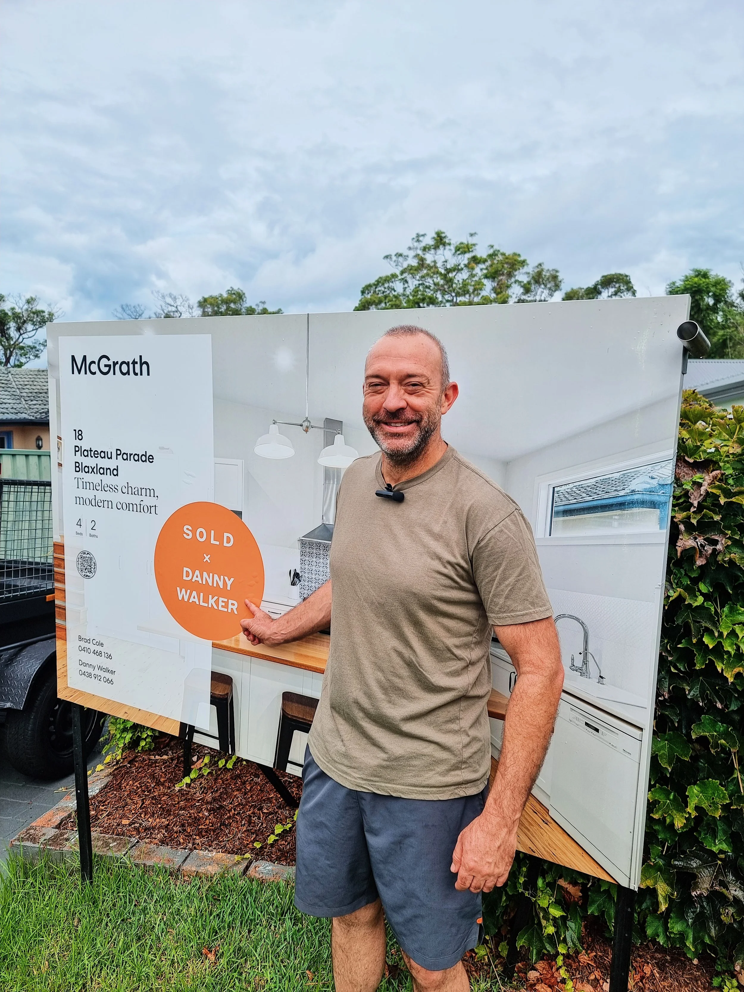 A man standing outdoors next to a large real estate sign, pointing at it and smiling. The sign features a photo of a modern kitchen interior with white cabinets and a window. The sign indicates the property is sold, with contact information for the r