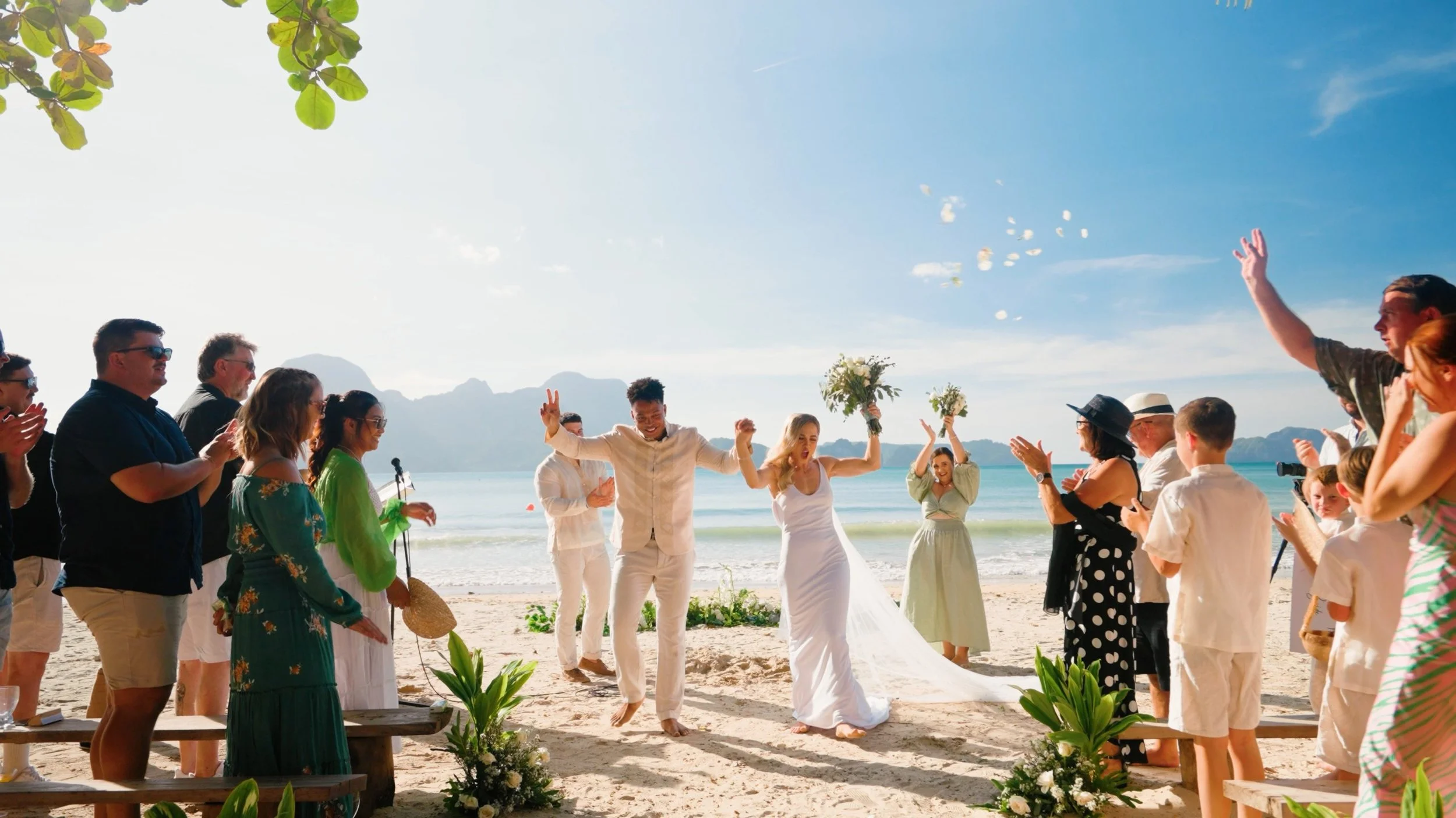 A beach wedding celebration with a bride and groom jumping together, surrounded by friends and family clapping and cheering on the sand, with mountains and ocean in the background on a sunny day.