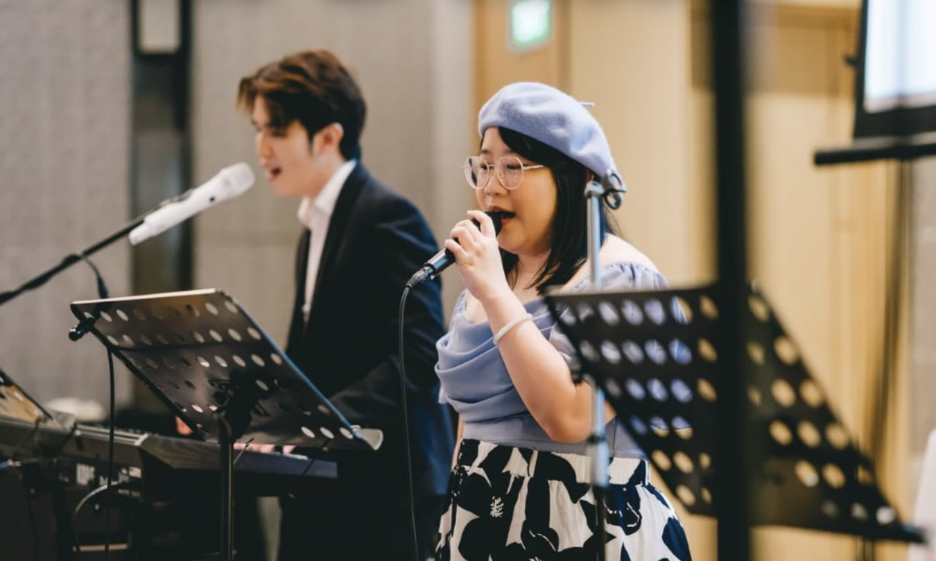 Two people singing into microphones during a musical performance in an indoor venue, with music stands and electronic keyboard equipment.