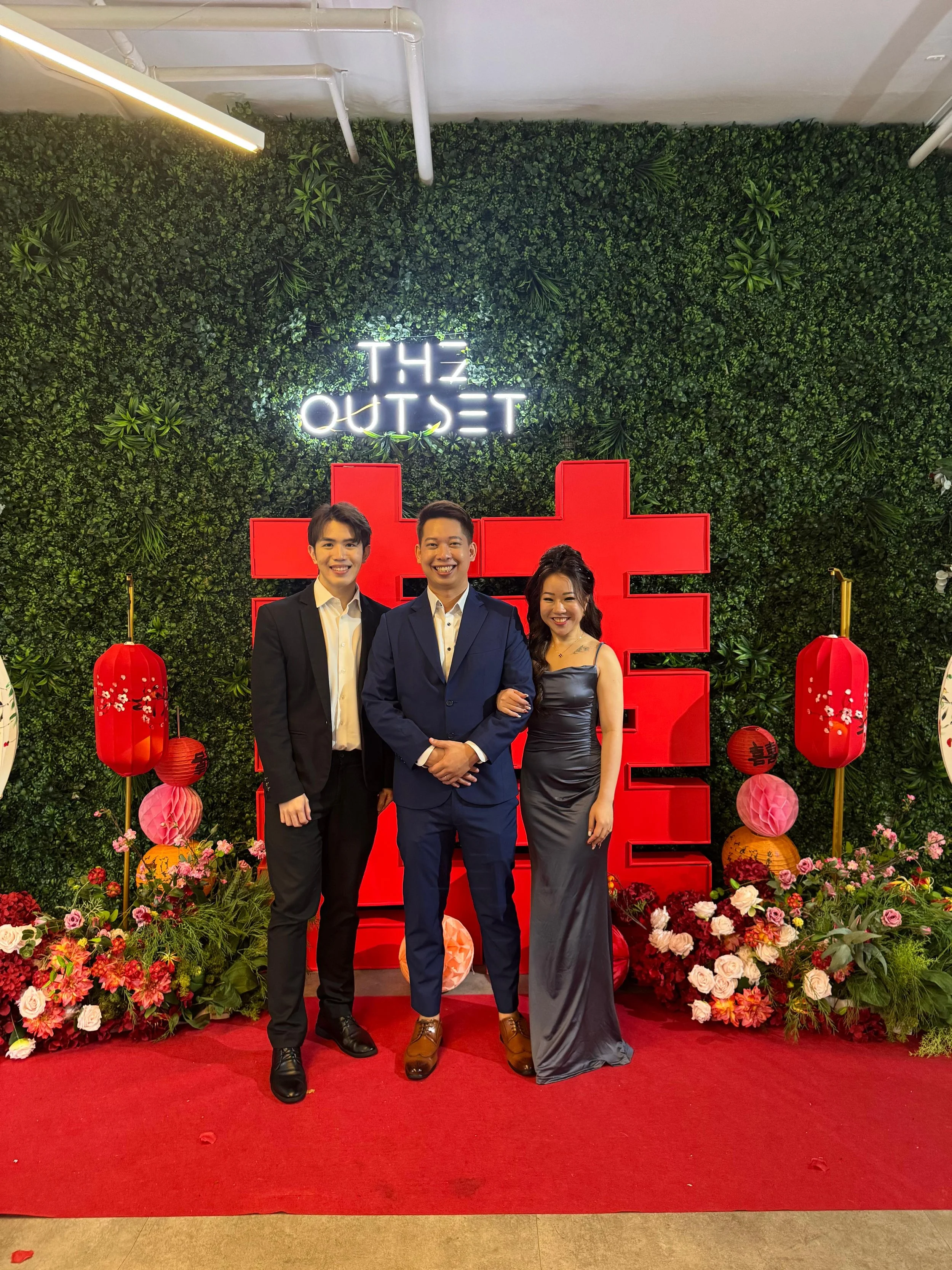Three people dressed in formal attire standing in front of a decorative backdrop with green foliage, red lanterns, flowers, and a neon sign that reads 'The Outset'.