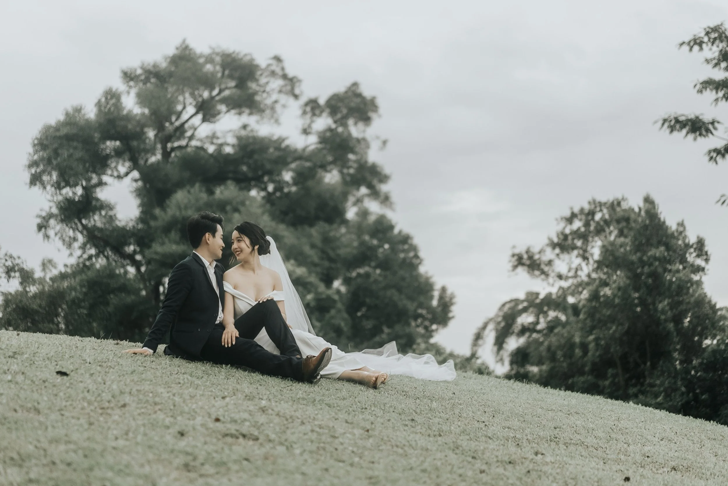 A couple, dressed in wedding attire, sitting on a grassy hill and smiling at each other with trees in the background.