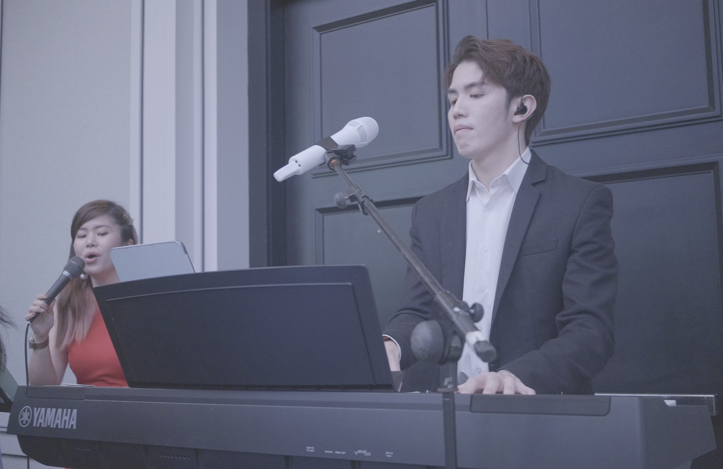 A young man in a suit is playing a Yamaha keyboard with microphone stands nearby, while a young woman in a red top sings into a microphone in the background.