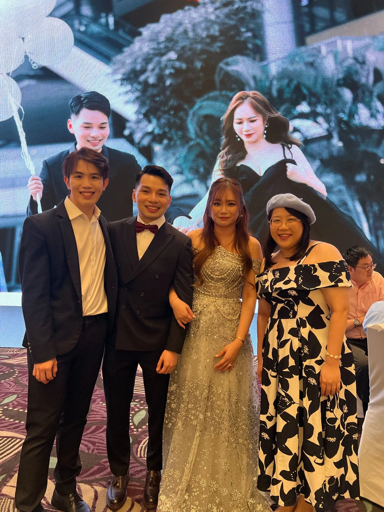 Four people in formal attire standing together in front of a large digital screen showing a photo of a couple. The group includes two men in suits, a woman in a silver gown, and a woman in a black and white off-shoulder dress with a grey beret. They are smiling and posing for the photo at a celebration or event.