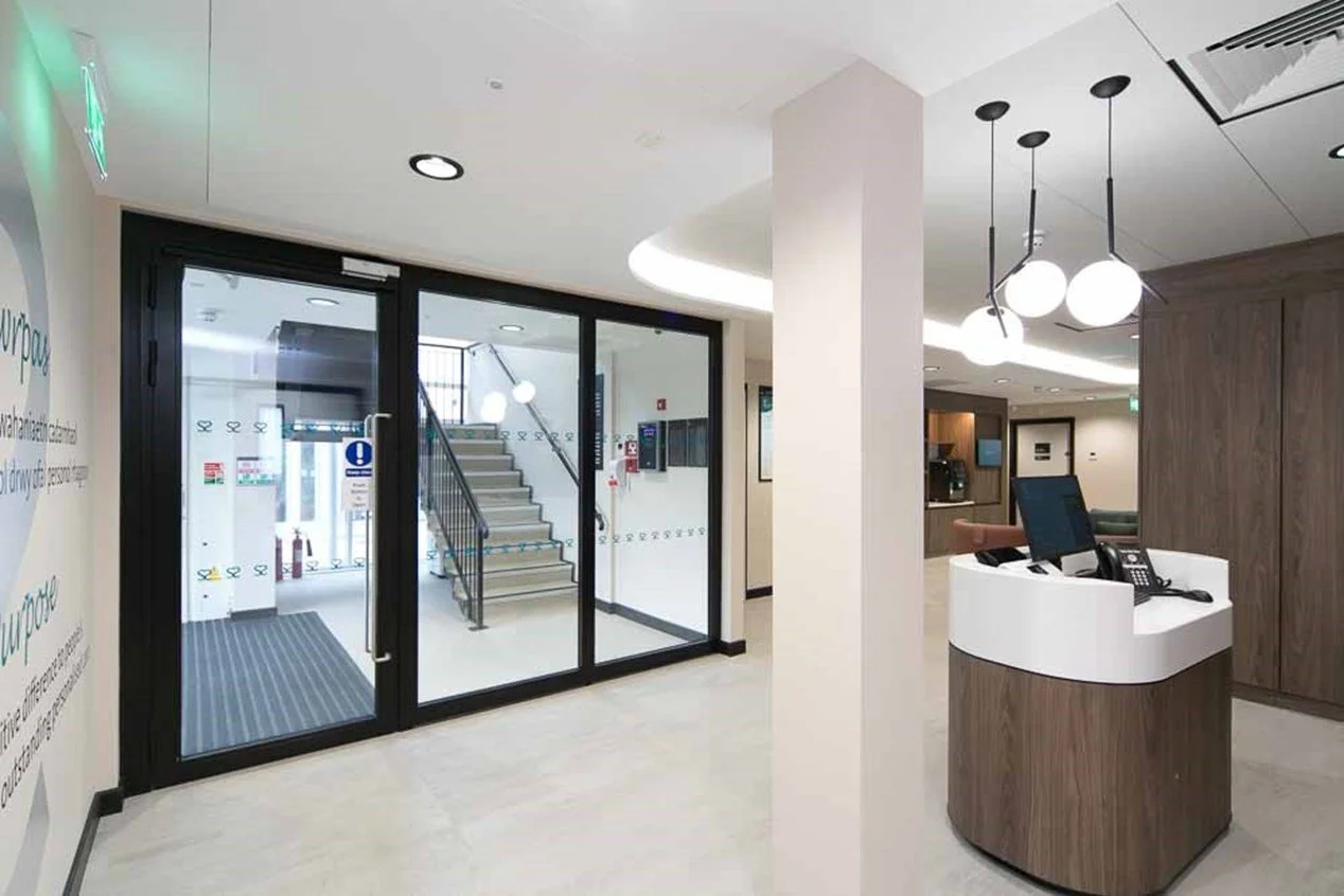 Modern reception with glass doors, a staircase, a reception desk with a computer and phone, and decorative hanging light fixtures